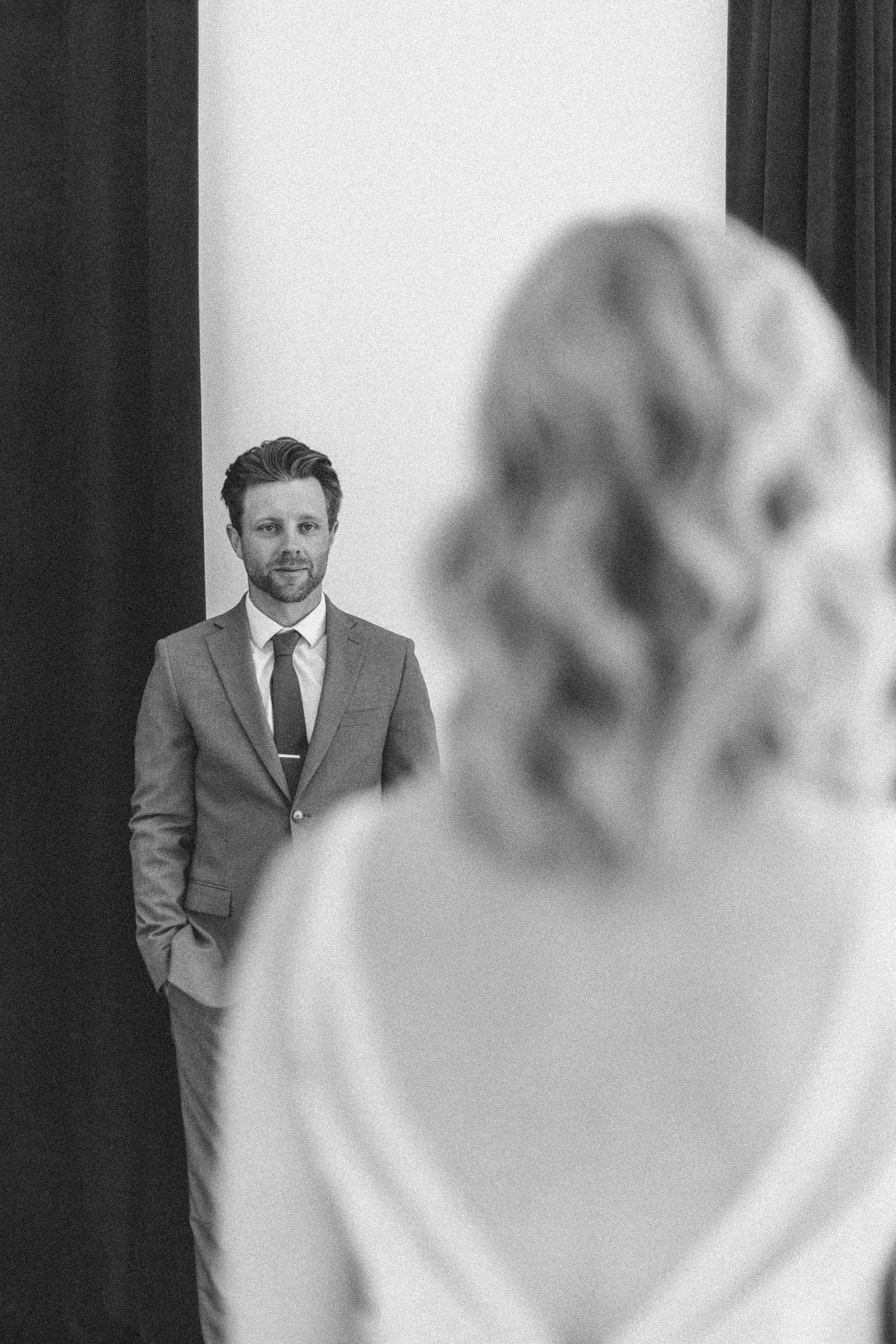Black and white photo of a groom in grey suit seeing bride in blurred foreground during first look moment.