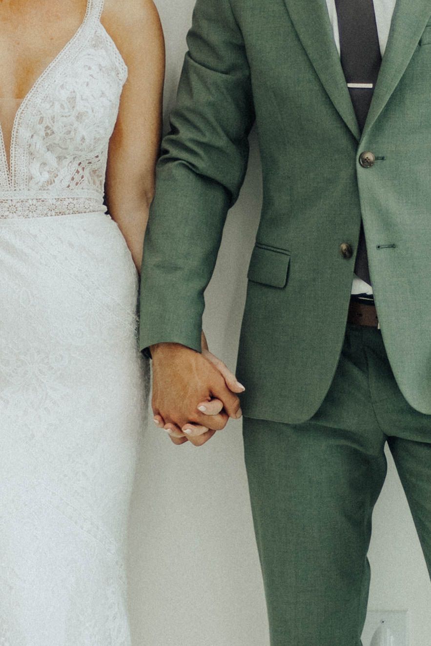 A close-up of a couple holding hands showing a white lace wedding dress and sage green suit details.