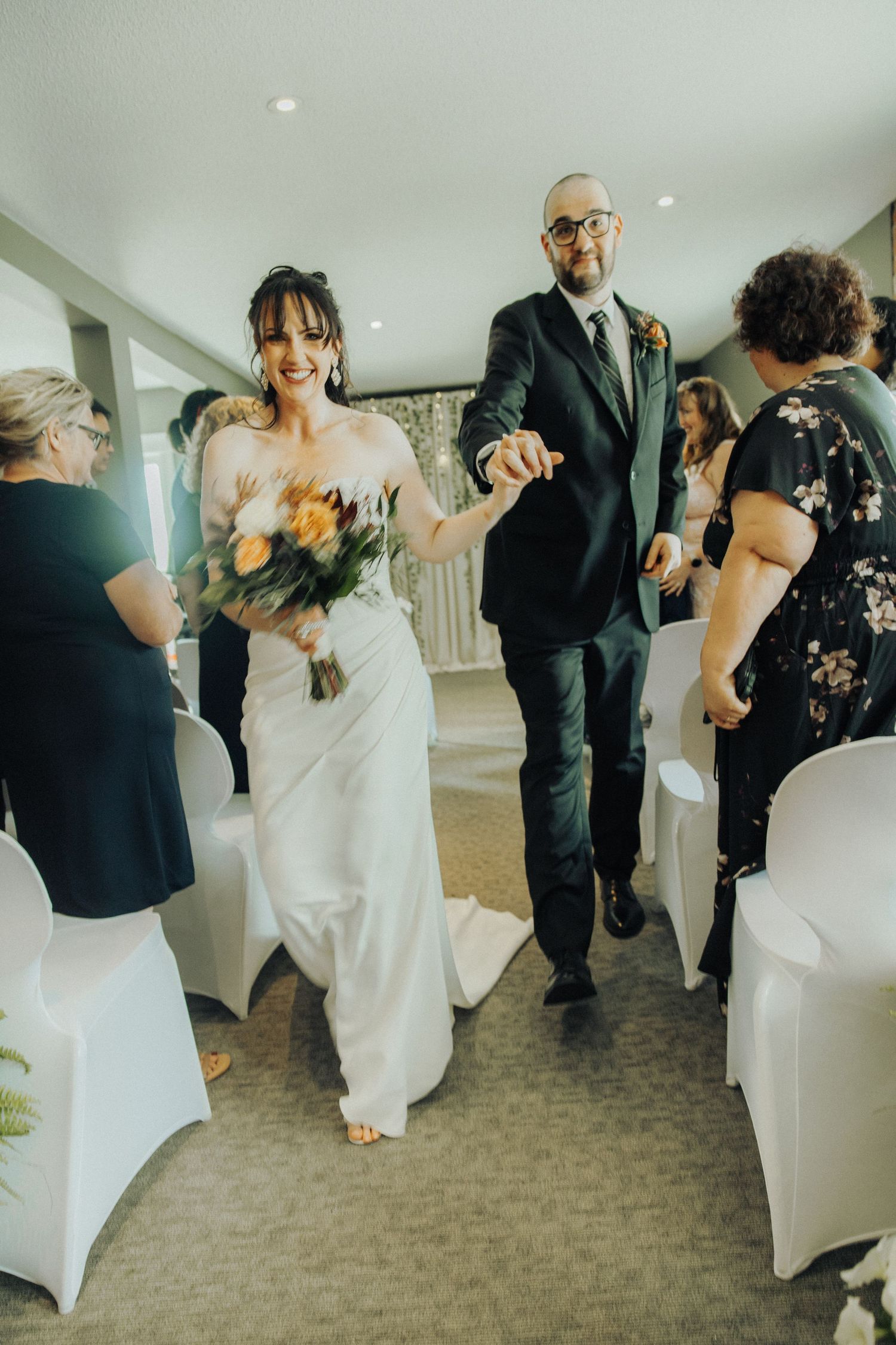 Smiling bride holding bouquet walks down aisle hand-in-hand with groom in dark suit at indoor wedding ceremony.