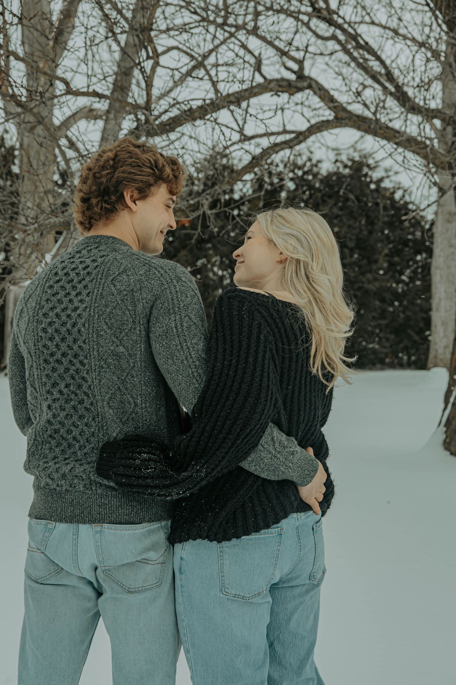 A couple in matching gray sweaters and jeans embrace while walking in a snowy winter landscape.