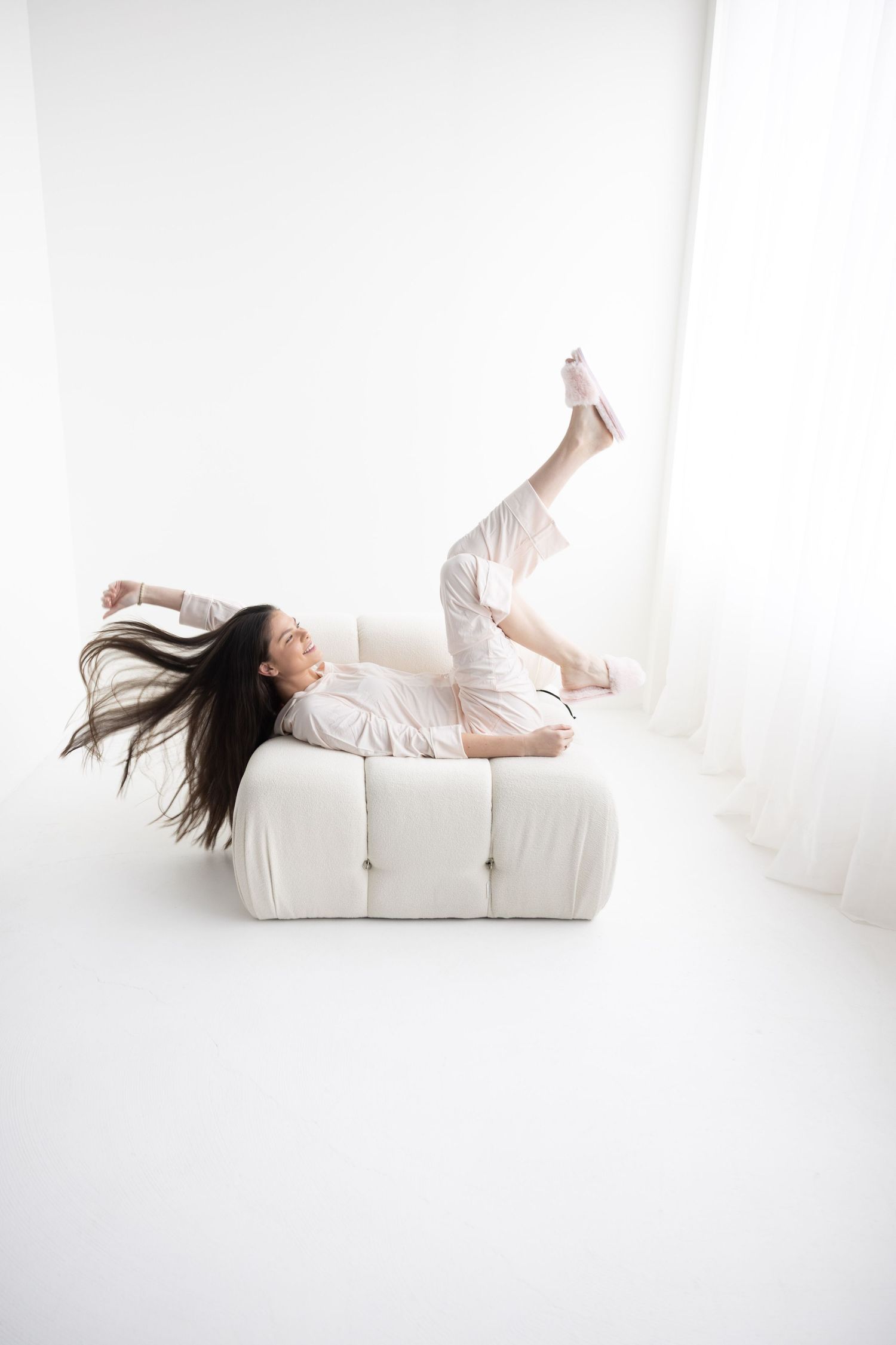 Woman in white outfit lounging dynamically on a white sofa with hair flowing in a bright white room.