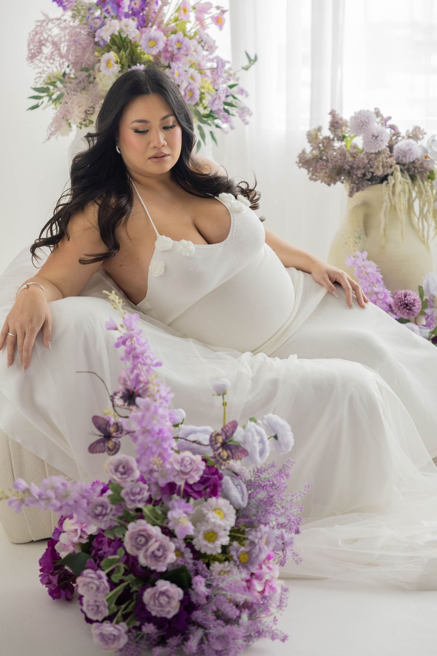 Elegant maternity photo with flowing white dress surrounded by purple and pink floral arrangements in bright setting.