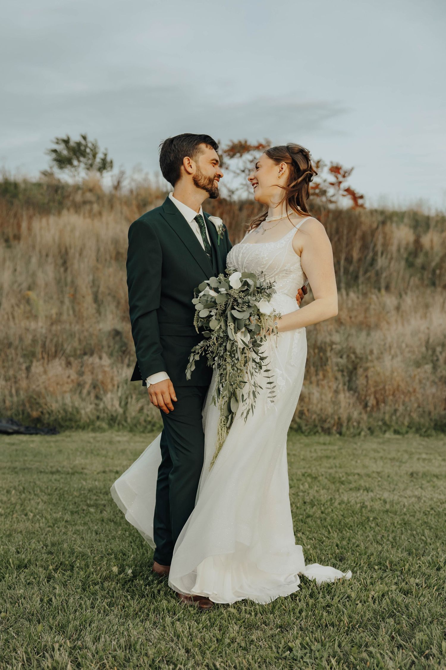 Romantic outdoor wedding portrait of couple in formal attire standing in a rustic field at sunset.