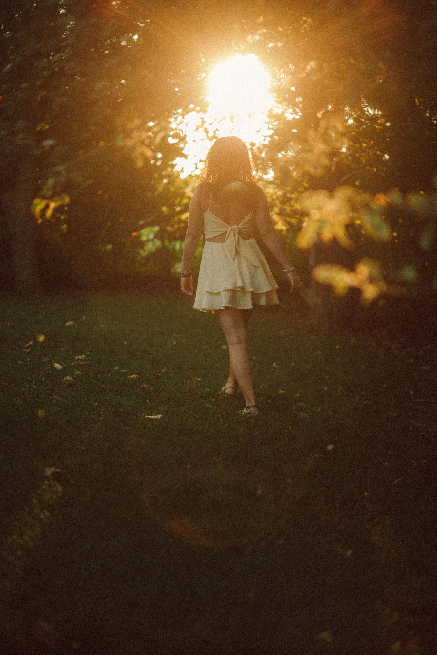 A young girl in a white tiered dress walks toward golden sunset light filtering through lush green trees in a garden.