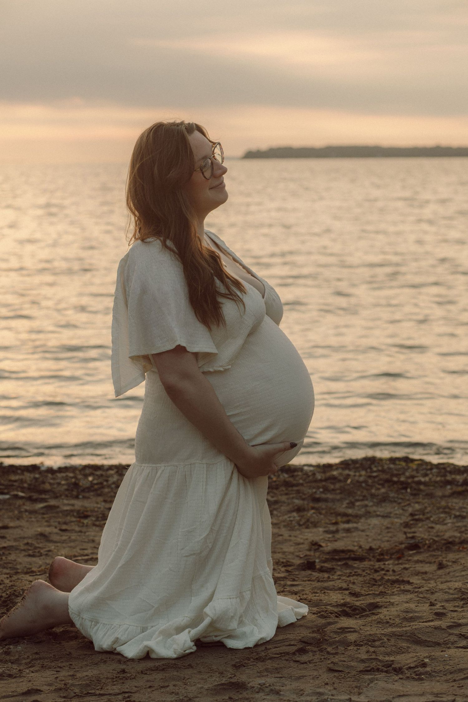 Pregnant woman in white dress kneeling on beach at sunset, cradling baby bump with serene ocean backdrop.