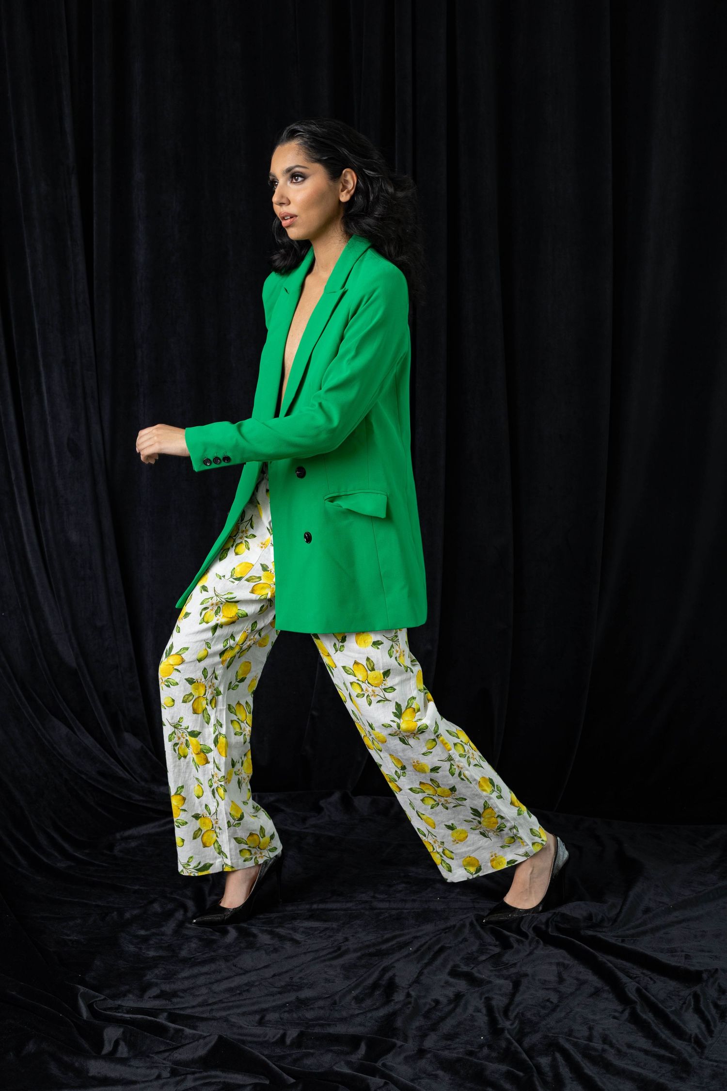 Fashion model strikes dynamic pose wearing vibrant green blazer with floral print pants against black backdrop.