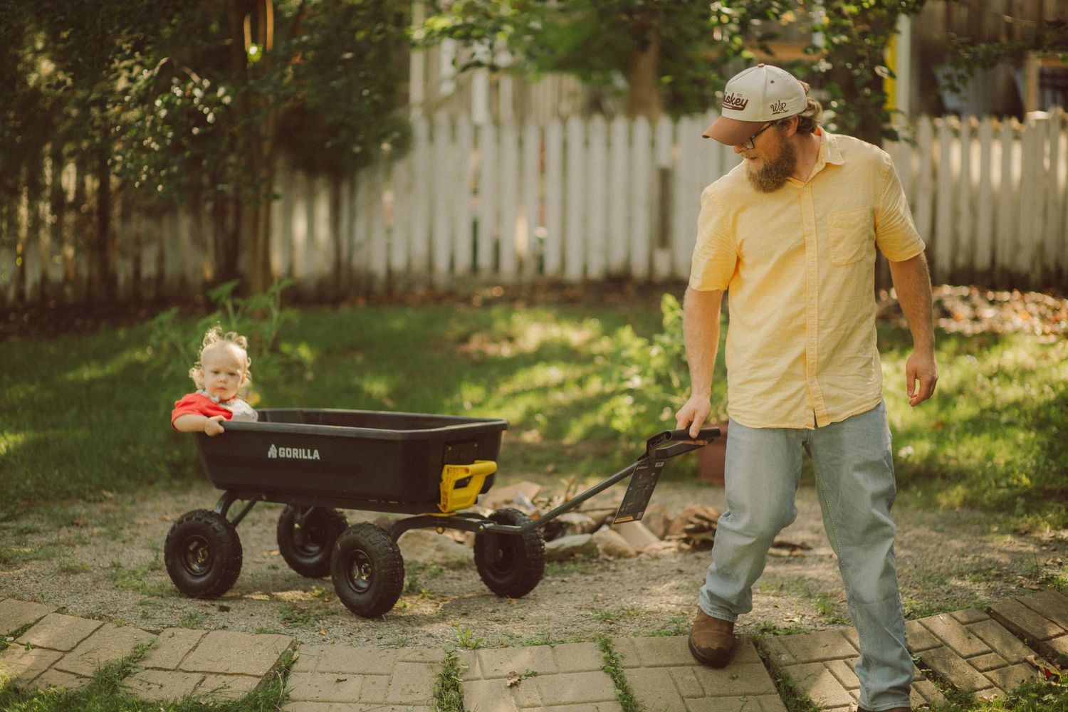 Bearded man in yellow shirt pulling a black garden wagon with a baby sitting inside on a brick patio.