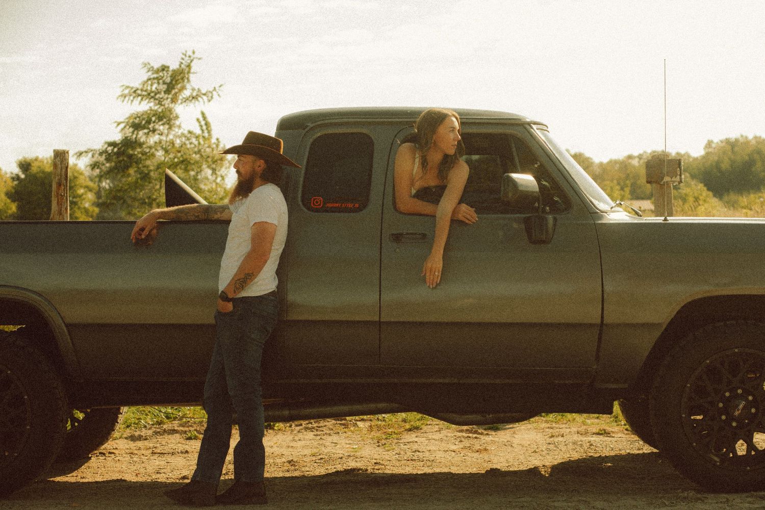 A man in a cowboy hat leans against a vintage green pickup truck while a woman leans out the window at sunset.