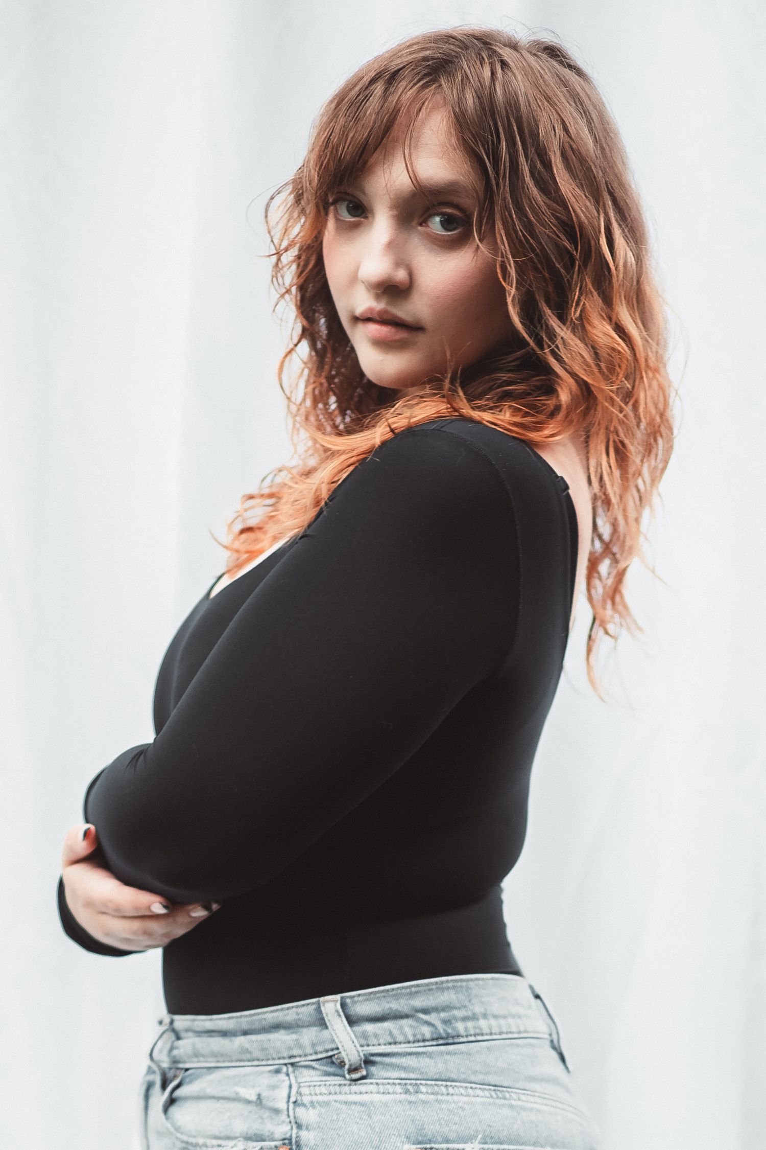 Young woman with wavy auburn hair in black long-sleeve top and jeans, arms crossed, posing against white background.