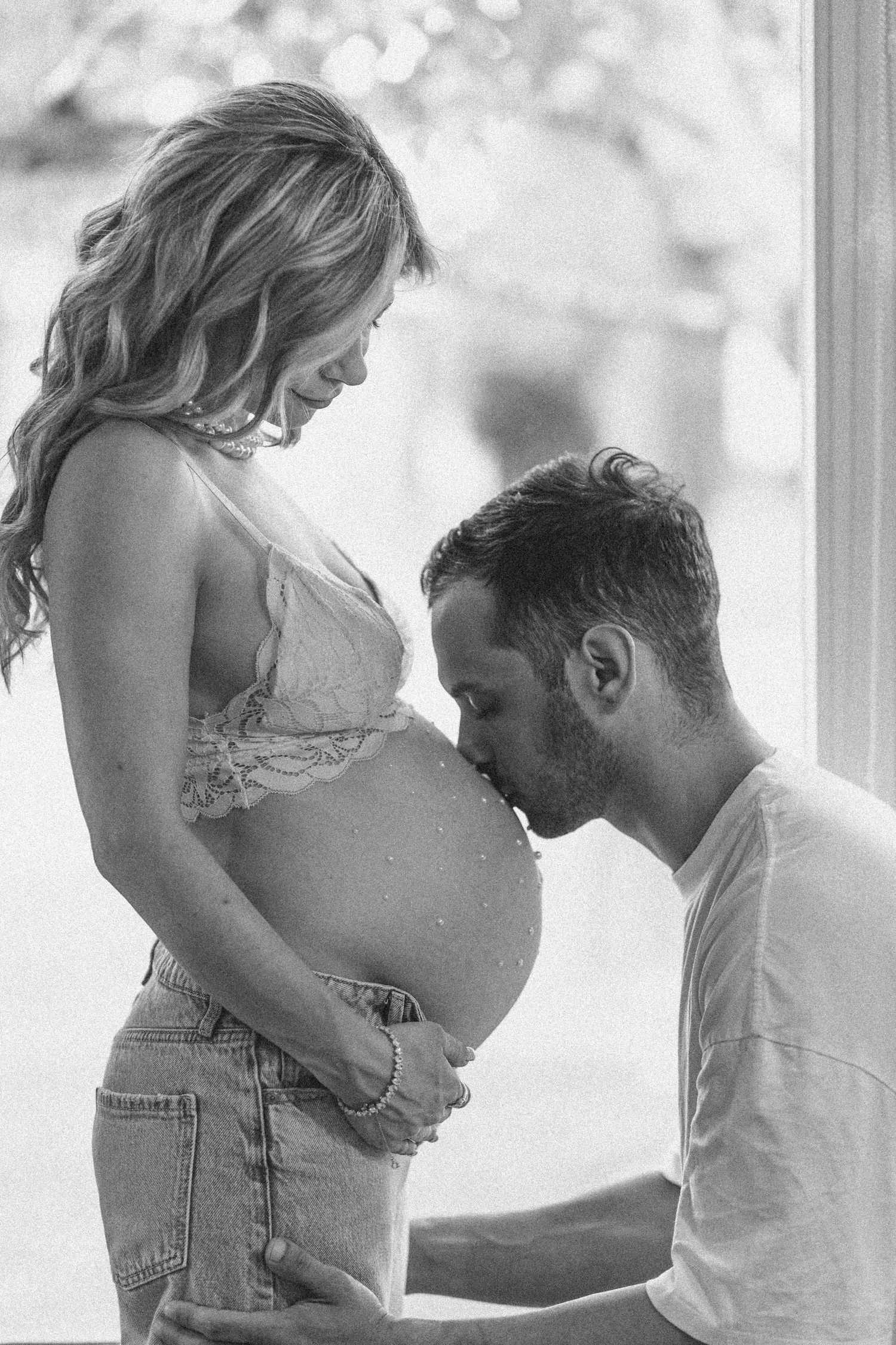 Black and white maternity photo of man kissing pregnant woman's belly near a bright window.