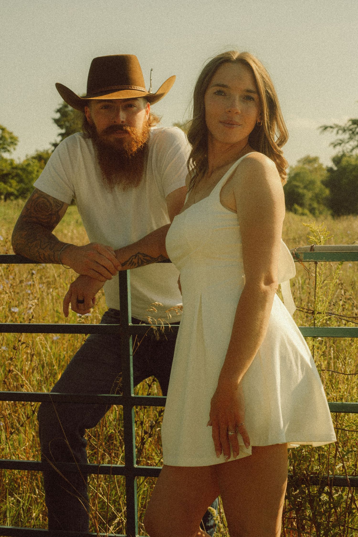 Tattooed bearded man in cowboy hat posing with young woman in white dress by a fence.