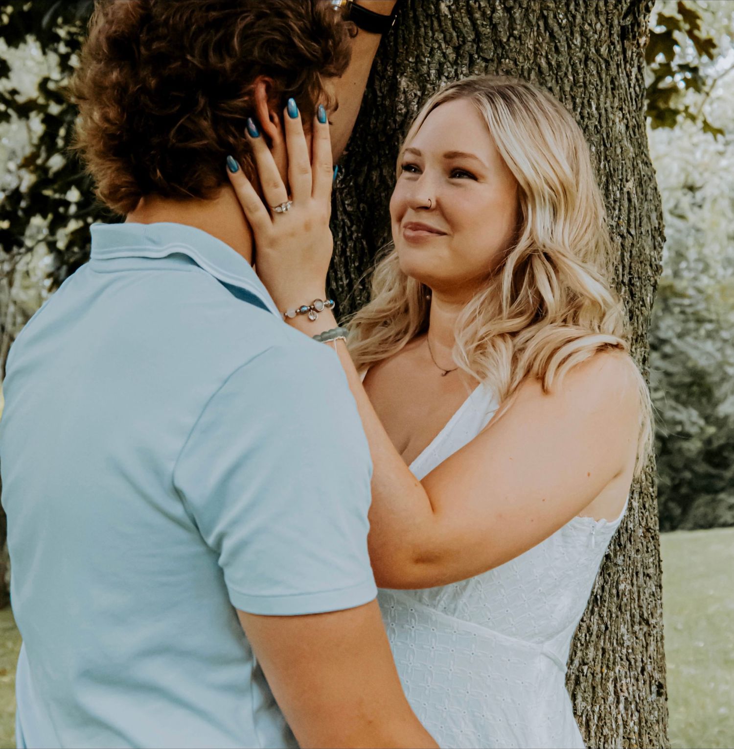 A couple shares a tender moment leaning against a tree trunk during a sunny outdoor photo session.
