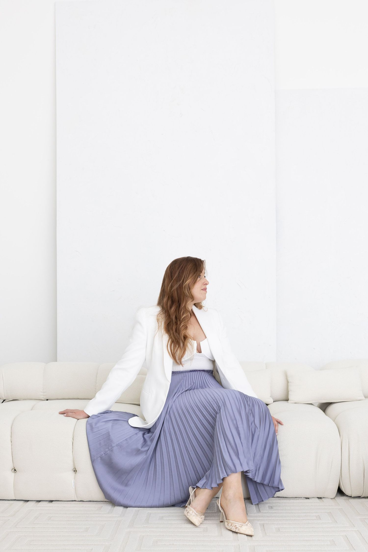Woman in white blazer and blue pleated midi skirt sitting on a cream sofa in a bright white room.