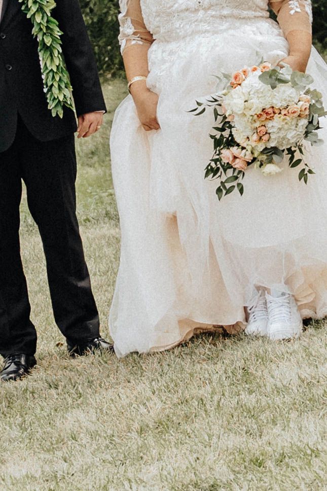 Wedding party members stand together on grass while holding elegant bouquets of peach and white flowers with greenery.