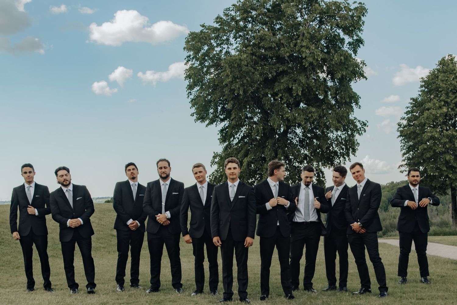 Groom and groomsmen in matching black suits pose outdoors on green grass with a large tree and blue sky behind them.