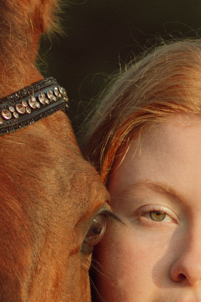 Close-up of a red-haired woman with green eyes resting her face against a chestnut horse wearing a decorative bridle.