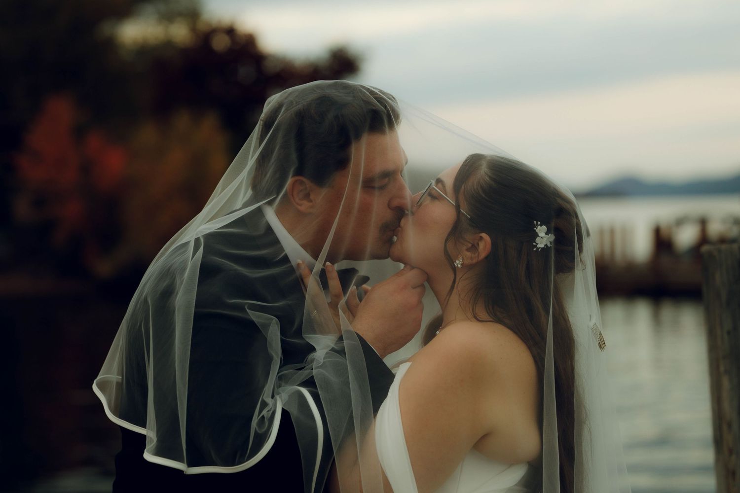 Romantic couple in wedding attire sharing a tender kiss under a flowing veil at sunset by the water.