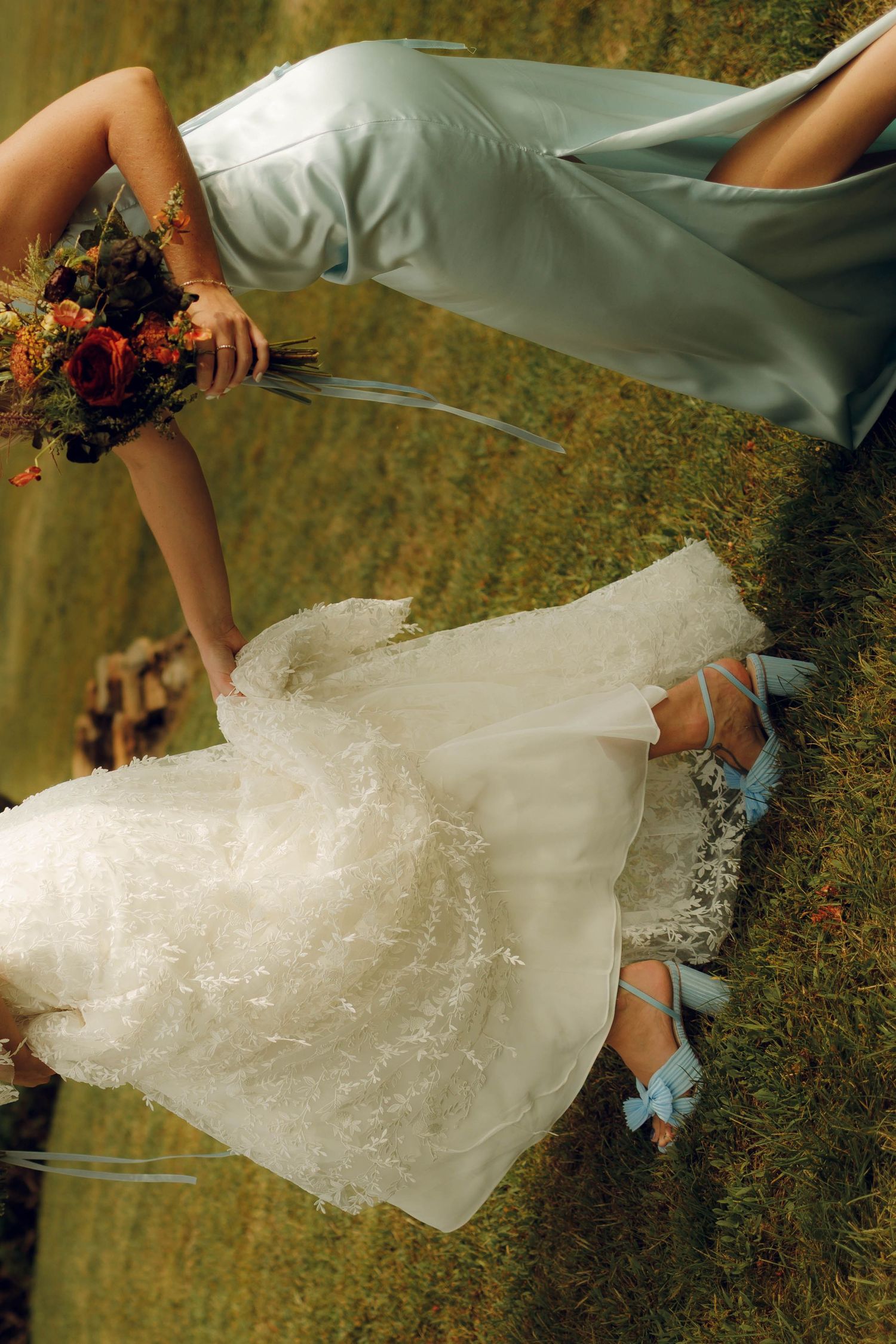 A flowing white wedding dress with delicate lace details is photographed against a grassy outdoor backdrop.