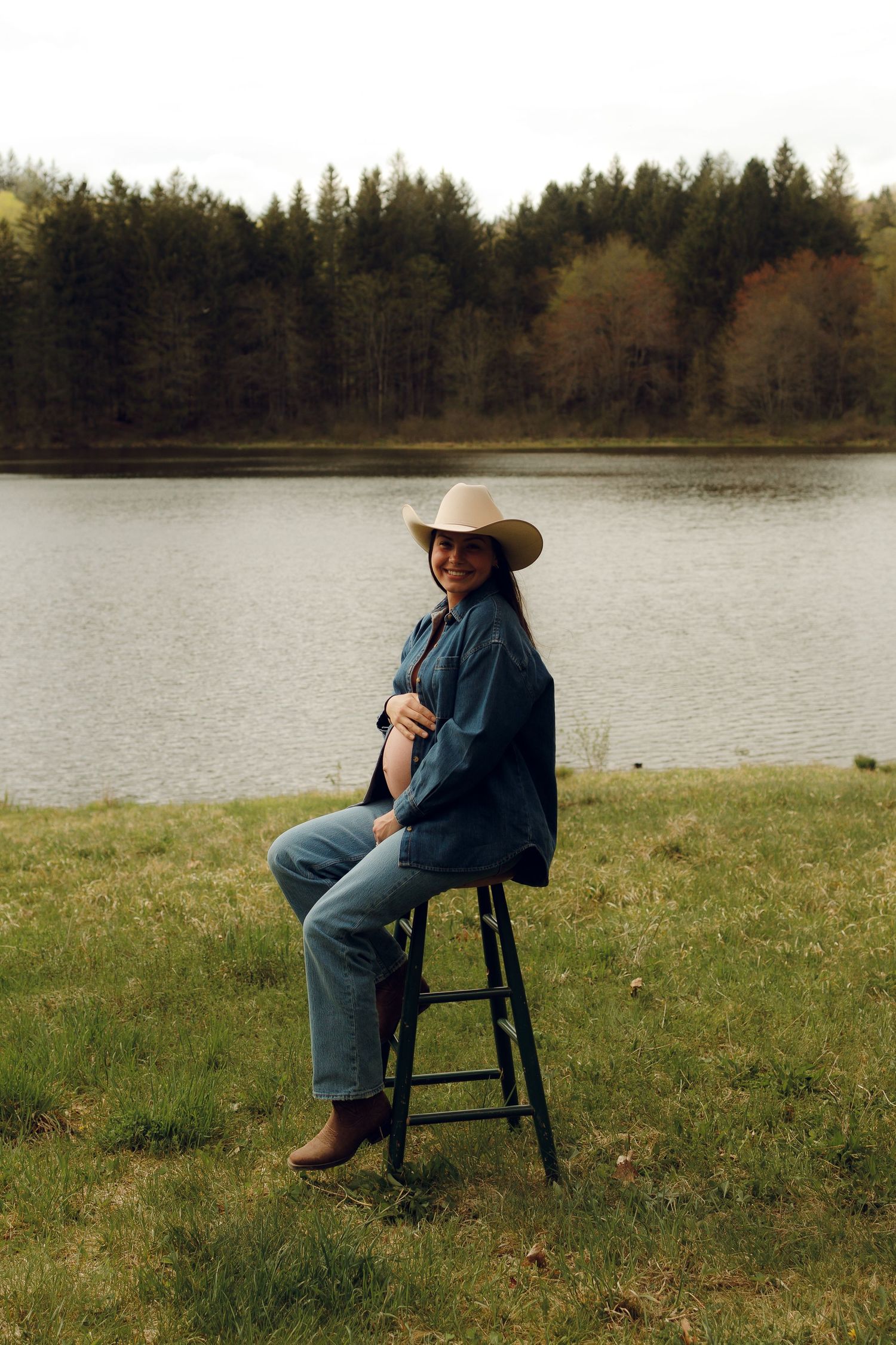 Person in denim and cowboy hat sits on stool by serene lake with evergreen forest backdrop on cloudy day.