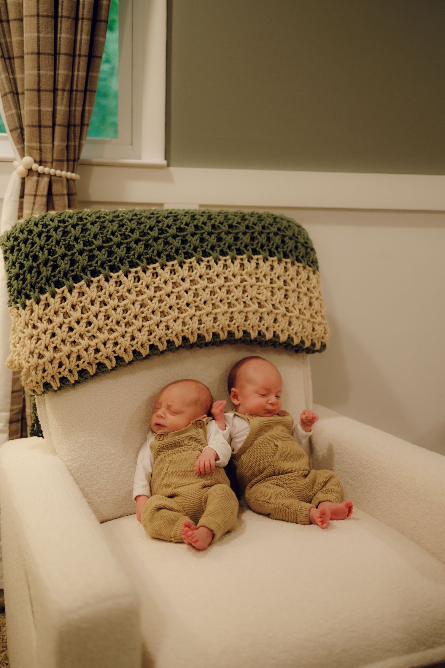 Two infants wearing tan outfits sleep side by side on a white armchair beneath a knitted green and beige blanket.
