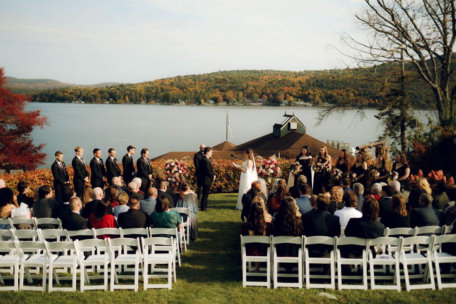 Panoramic view of an outdoor wedding ceremony setup with white chairs overlooking a scenic autumn lake and mountains.