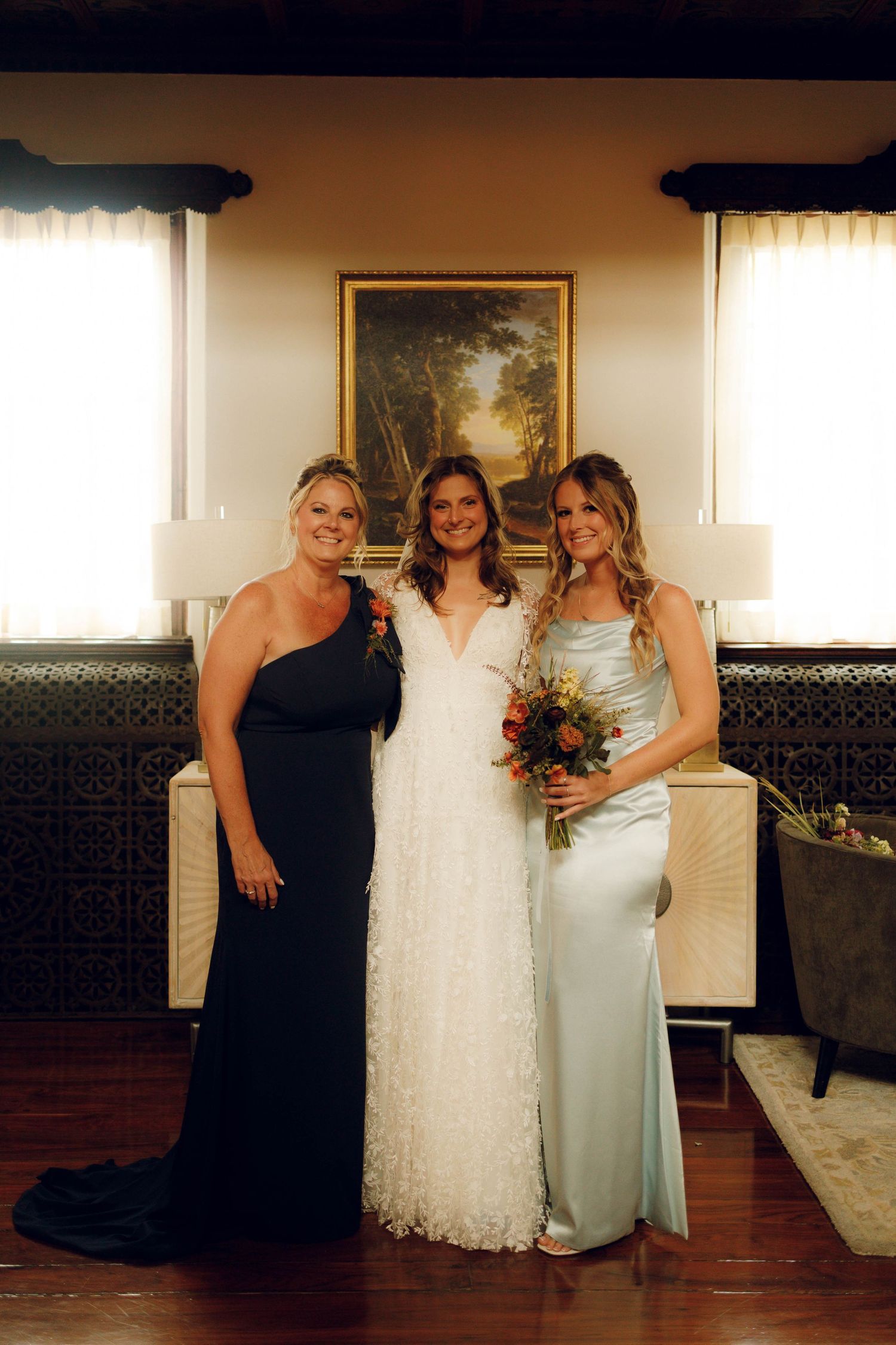 Three women in formal dresses pose together at an indoor wedding celebration with a painting on the wall behind them.