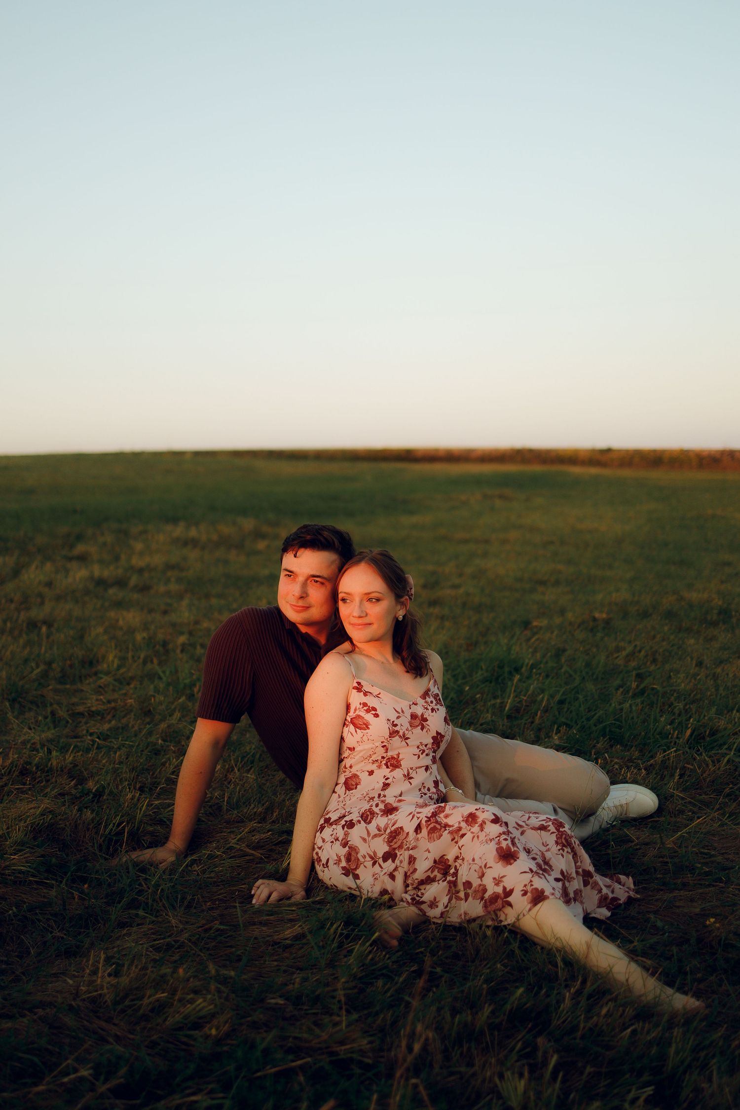 A couple relaxes together in a grassy field at sunset, creating a romantic silhouette against the horizon.
