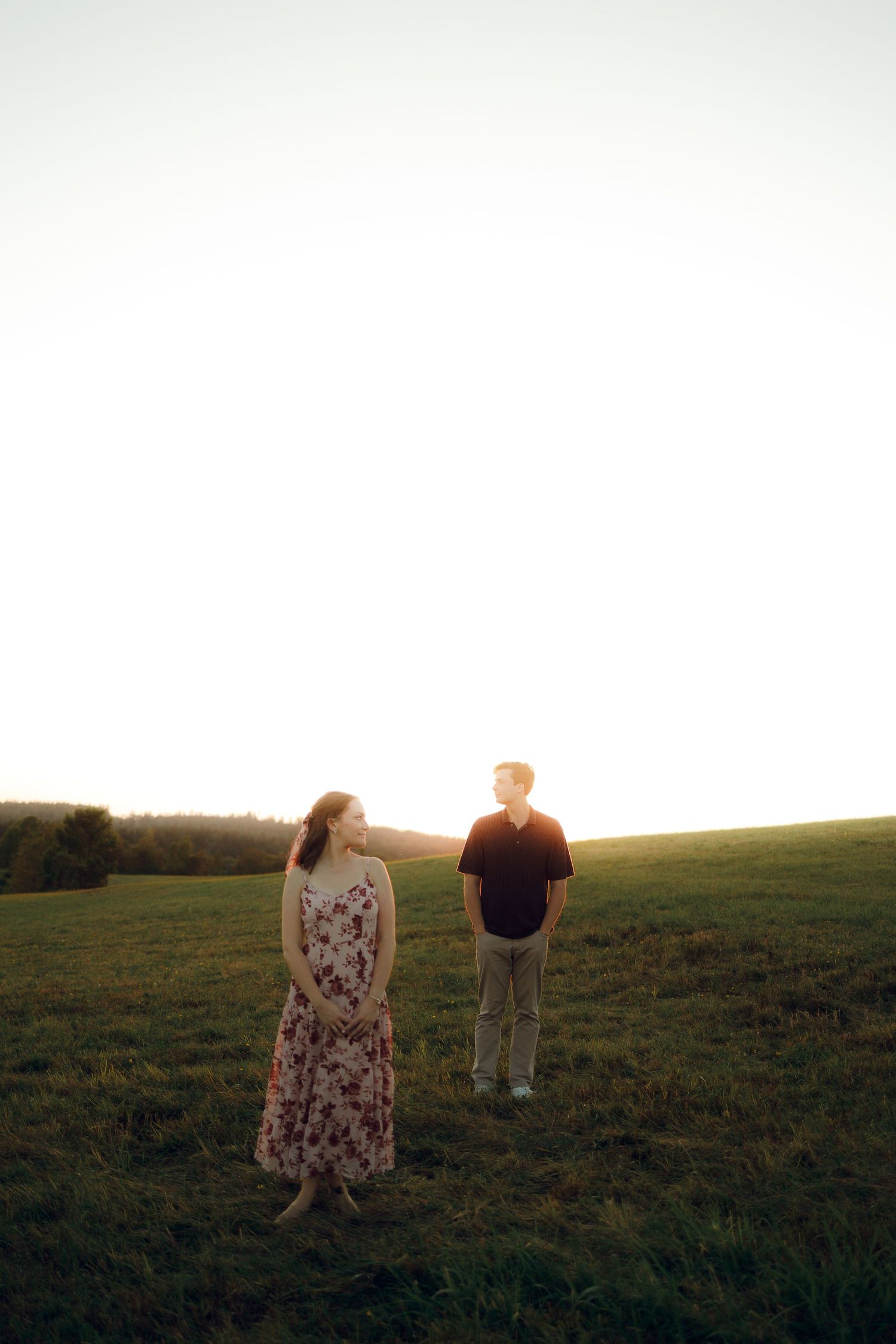 Two people stand together in a grassy field at golden hour as the sun sets over the horizon.