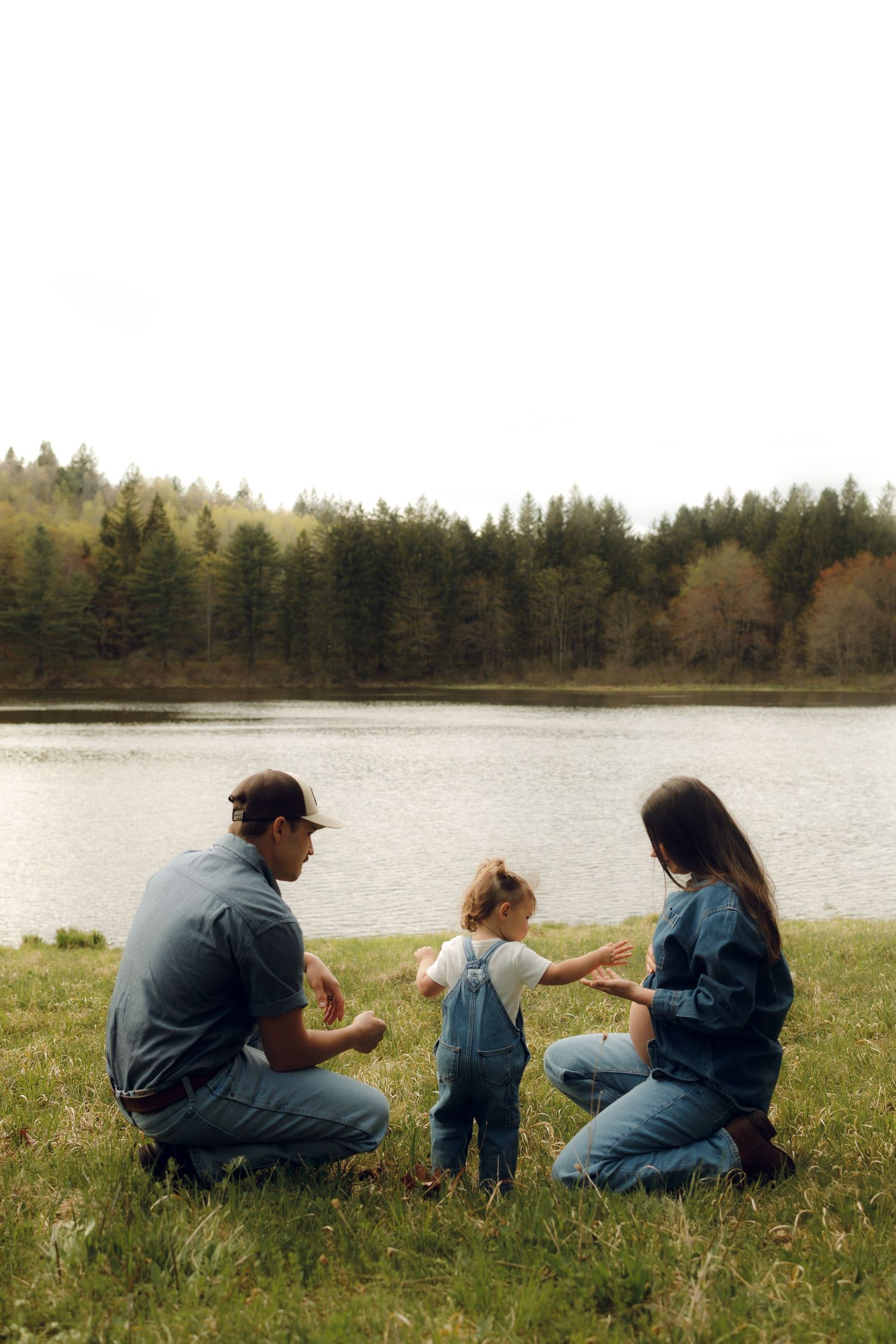 People crouched on grassy lakeside with pine forest and calm water in background on overcast day.