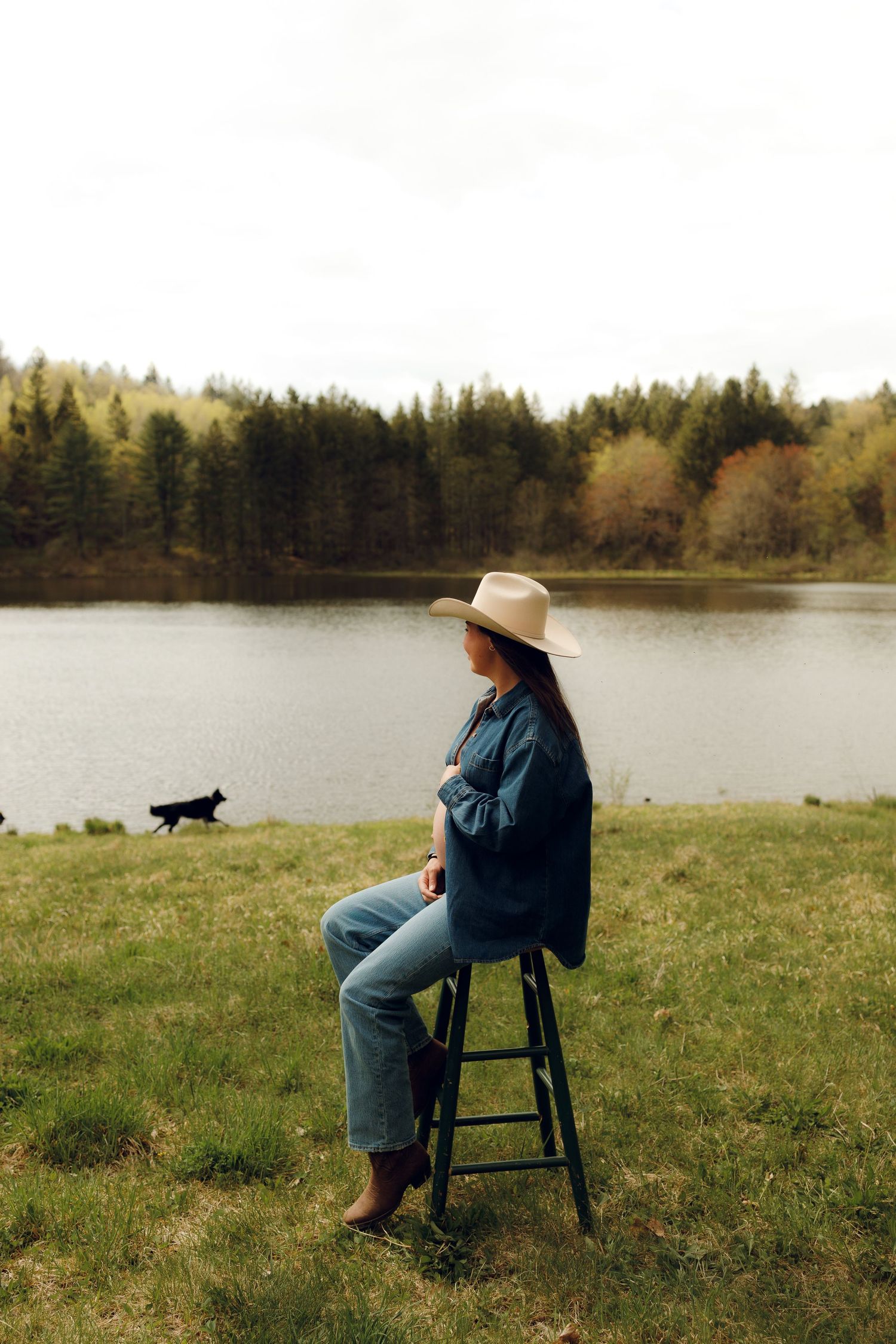 Person in denim jacket and wide-brimmed hat sits on stool by lake while small dog plays in background on grassy shore.