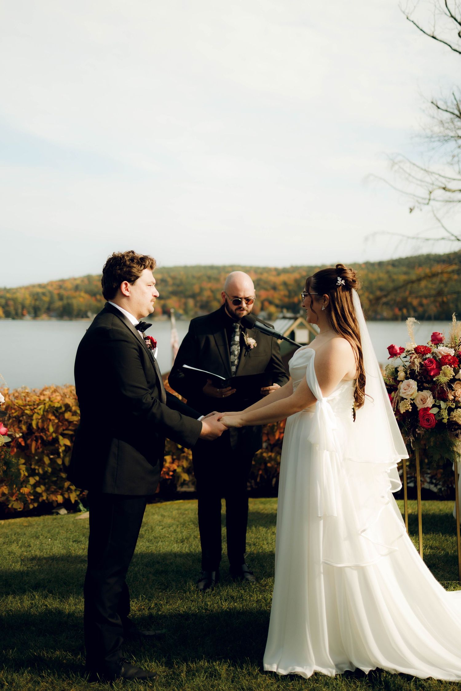 A romantic outdoor wedding ceremony takes place with a scenic lake and autumn foliage in the background.
