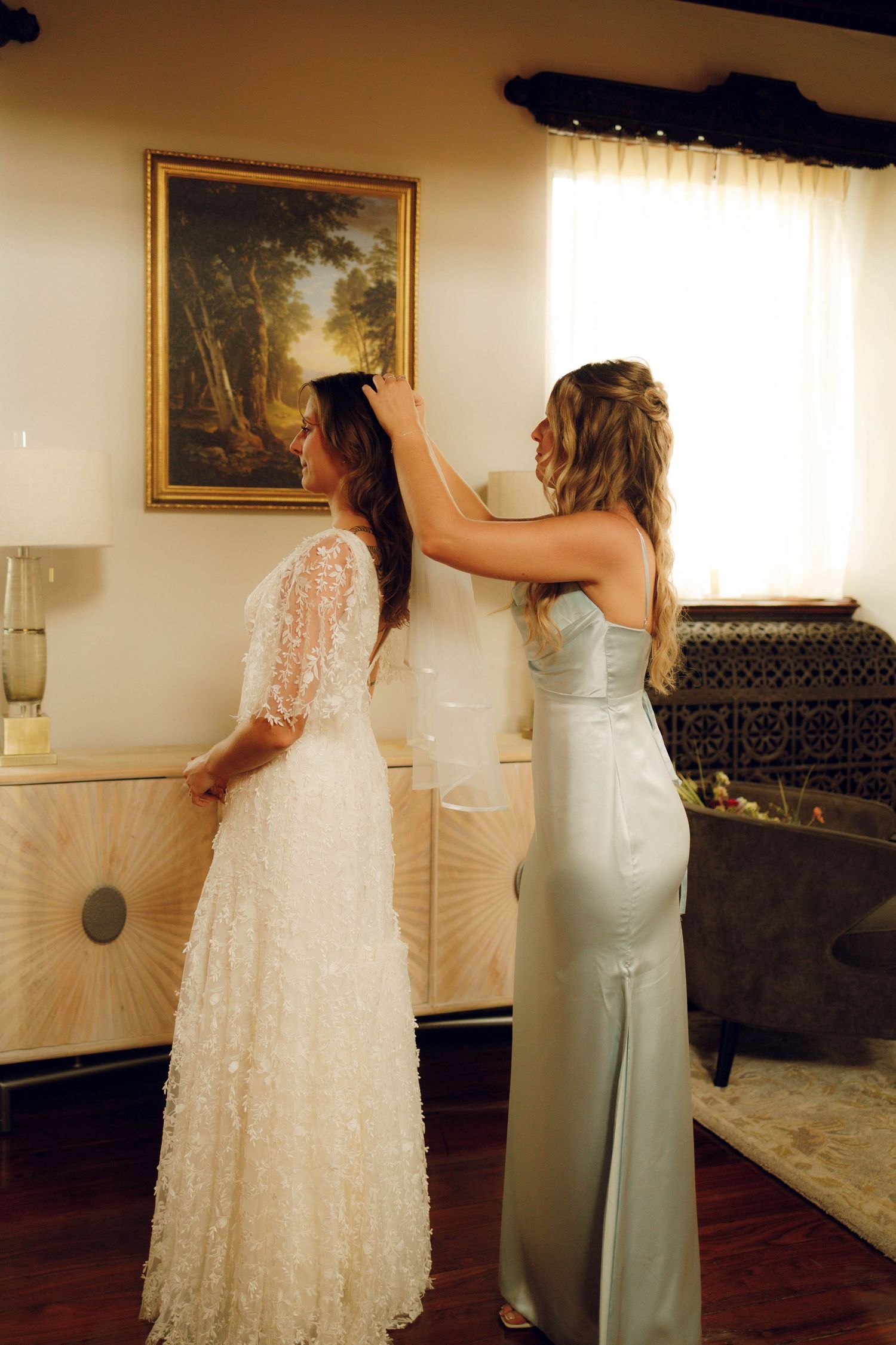 Two women in formal dresses stand together during a wedding preparation, with one adjusting the other's veil.
