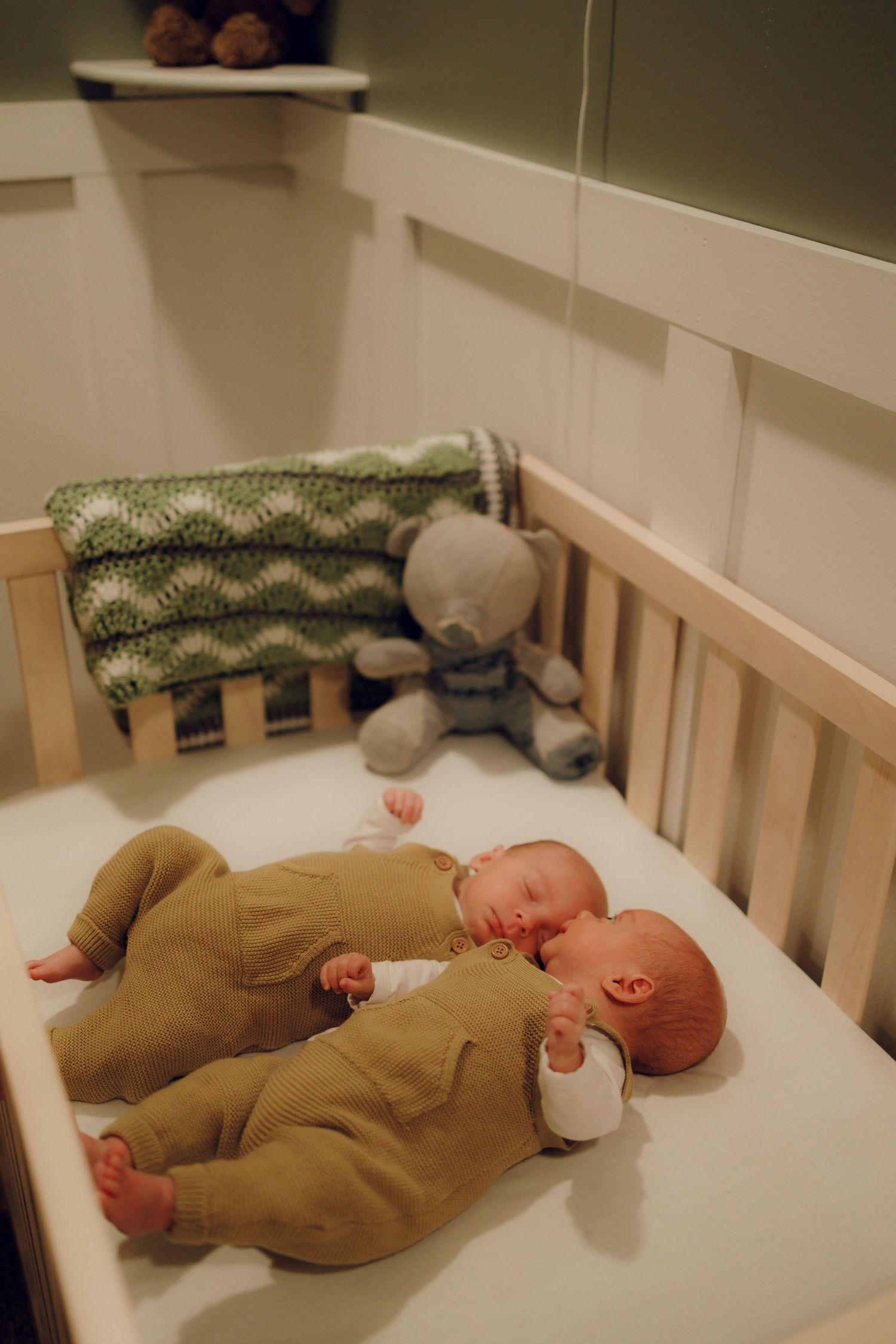 Two infants sleeping peacefully in a wooden crib with a teddy bear and patterned blanket in the corner.