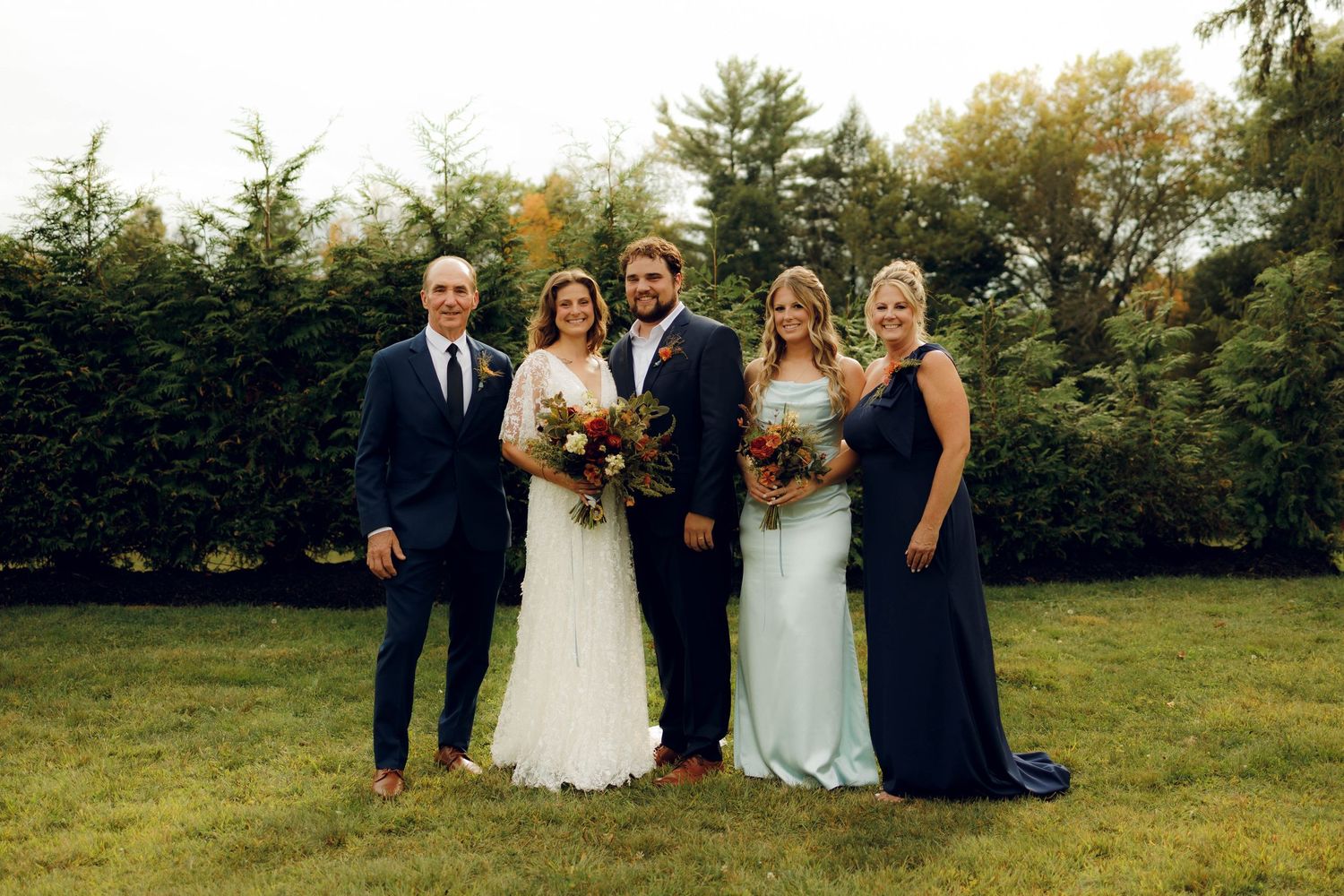 A wedding party poses together outdoors on a grassy hillside surrounded by autumn trees.