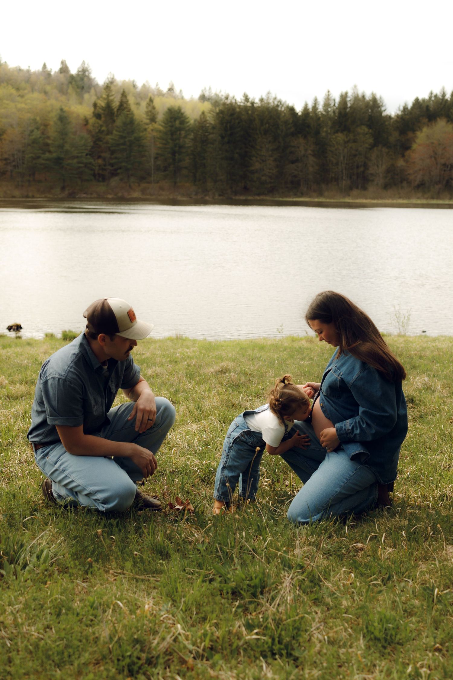 Family enjoying a peaceful moment together on grassy shore by lake with evergreen forest in background.