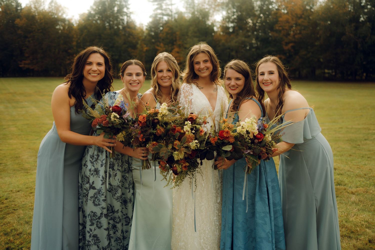 A bridal party wearing various shades of blue dresses holds colorful autumn bouquets in a sunlit field at sunset.