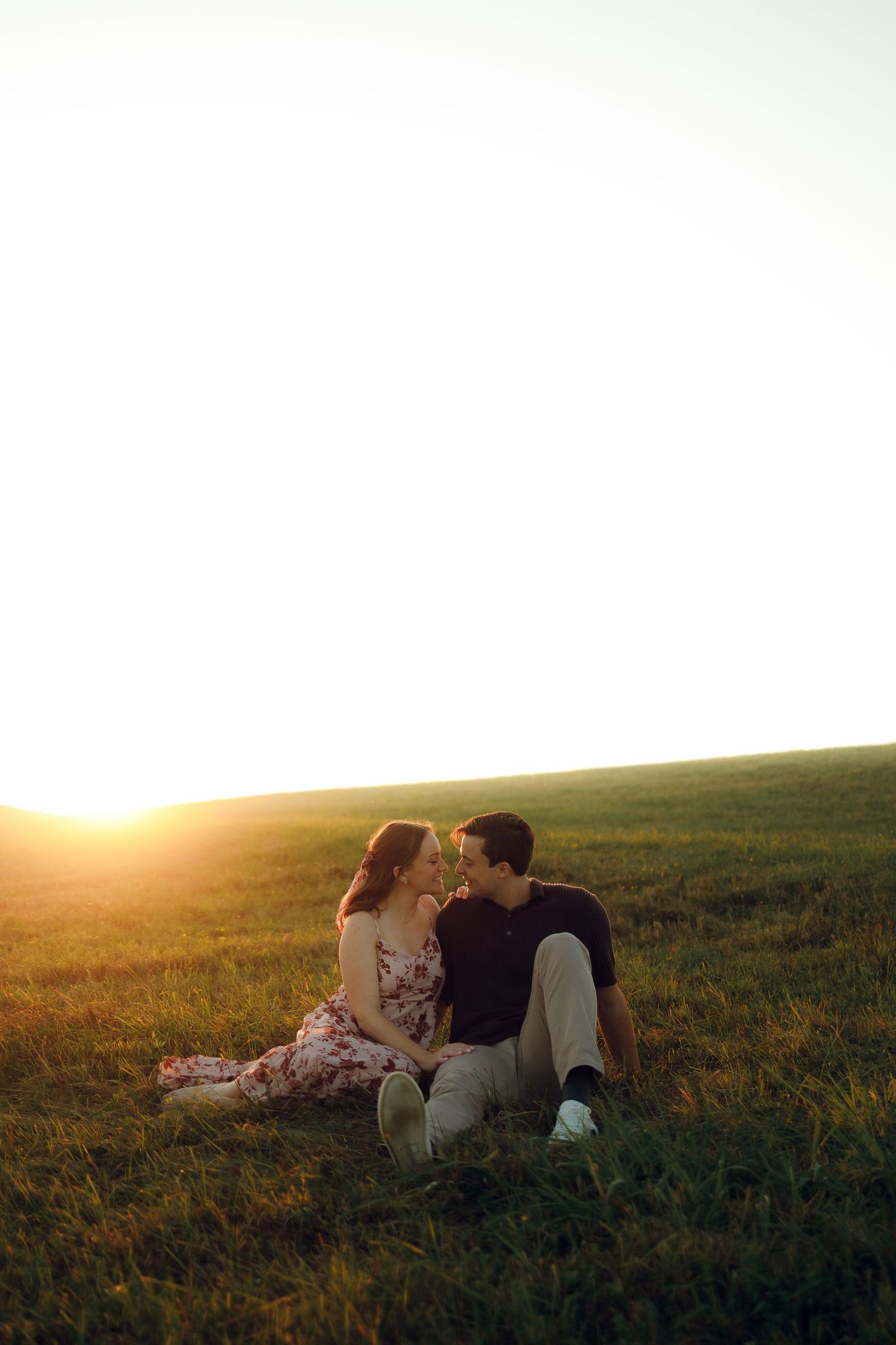 A couple sits together in a grassy meadow during golden hour sunset creating a romantic silhouette.