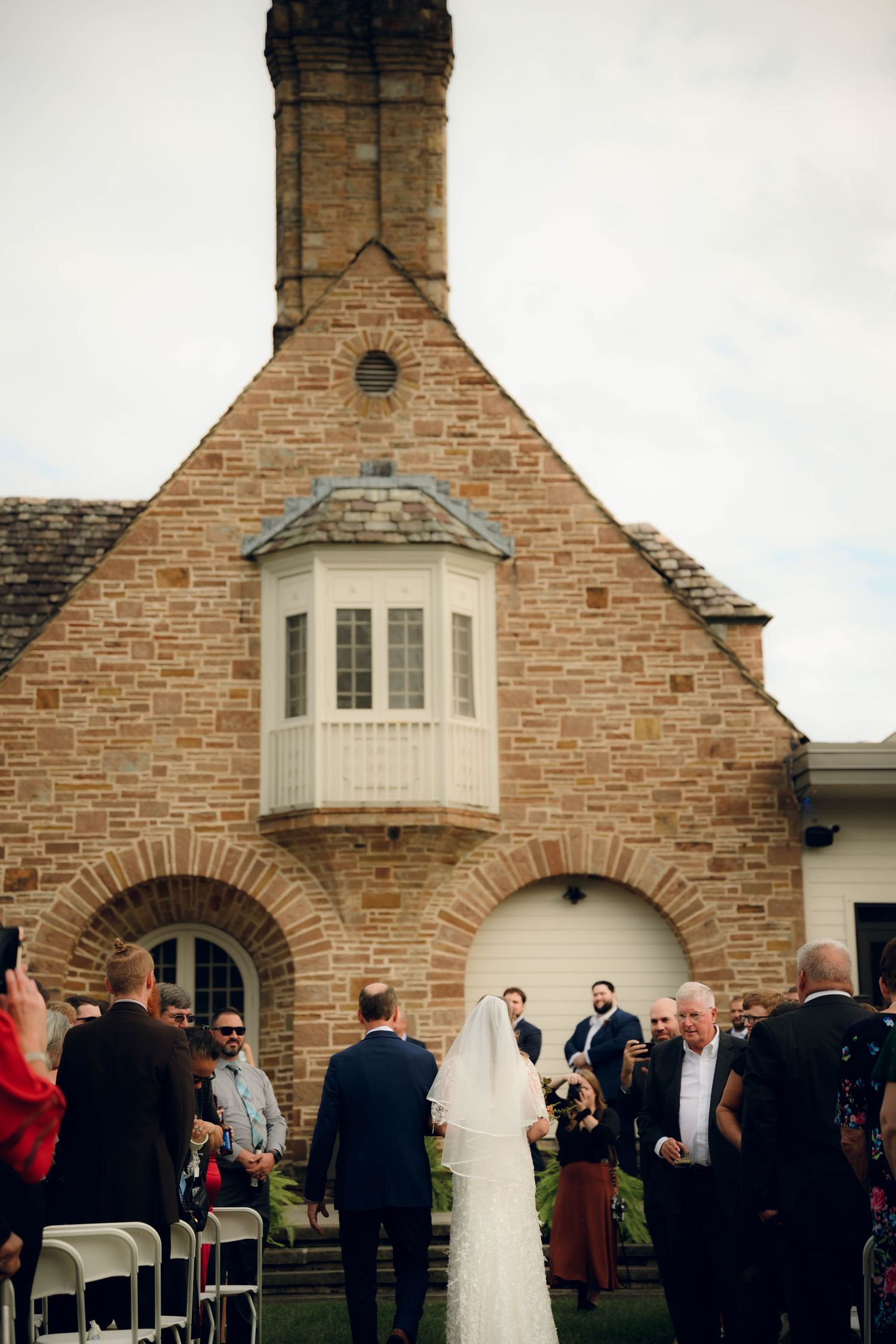 Wedding ceremony taking place outside a stone church building with distinctive bay window and arched entryways.