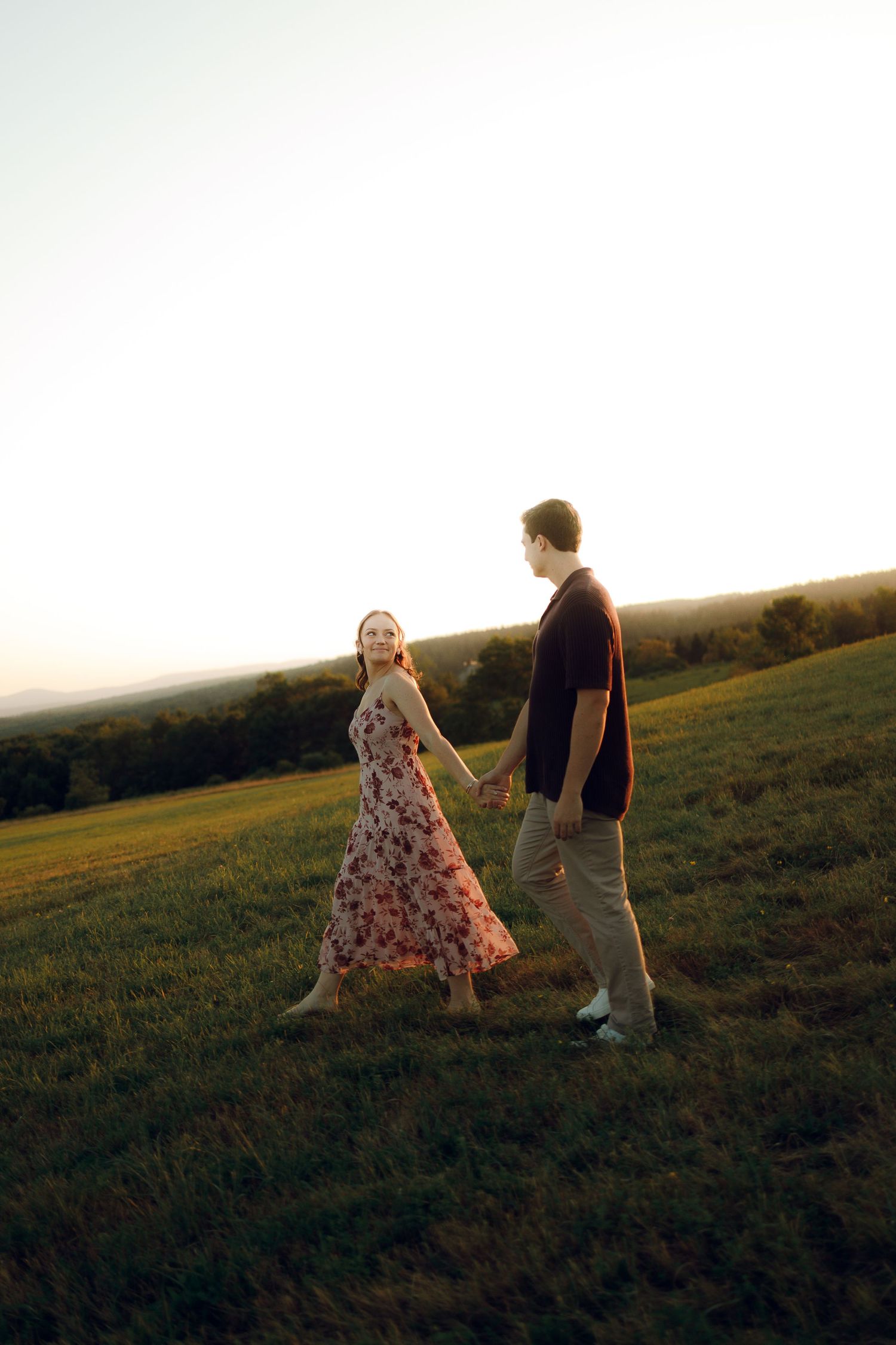 A couple walks hand in hand through a grassy hillside at sunset, with her floral dress flowing in the breeze.