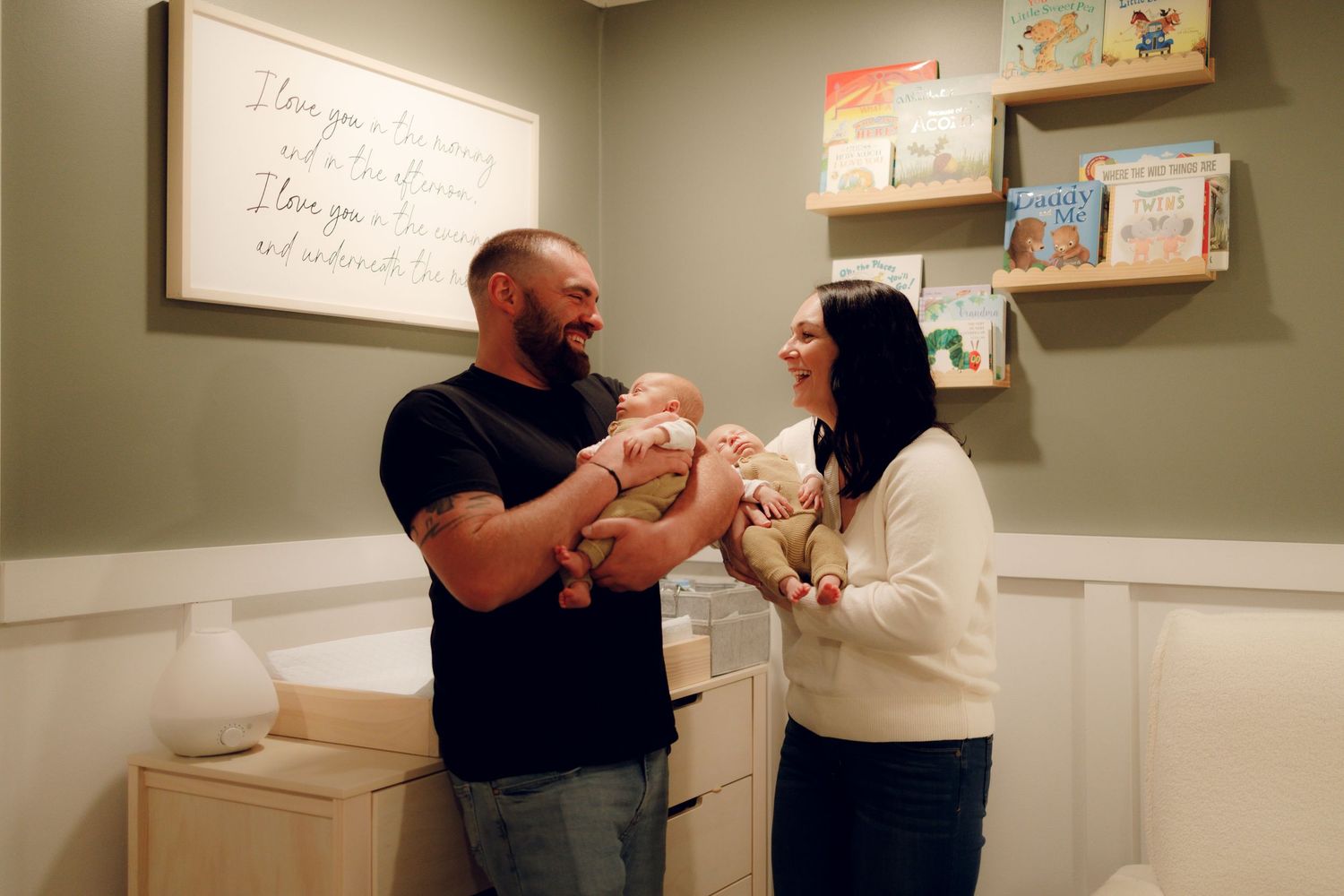 New parents share a tender moment with their newborn baby in a cozy nursery with sage green walls and floating shelves.