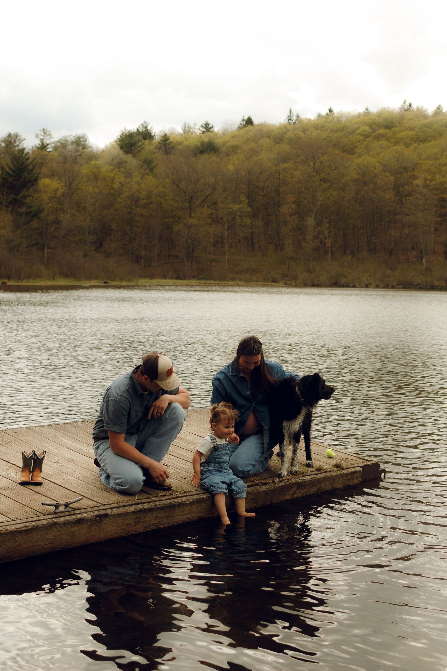 Family and dog spending time together on a wooden dock by a peaceful lake surrounded by wooded hills on an overcast day.