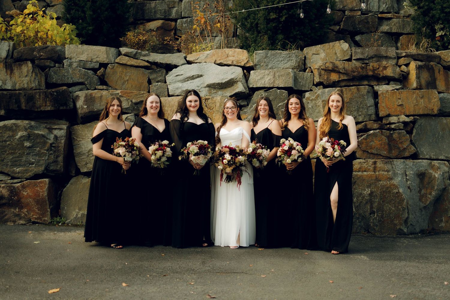 Wedding party in black bridesmaid dresses and white bridal gown pose together against stone wall backdrop.