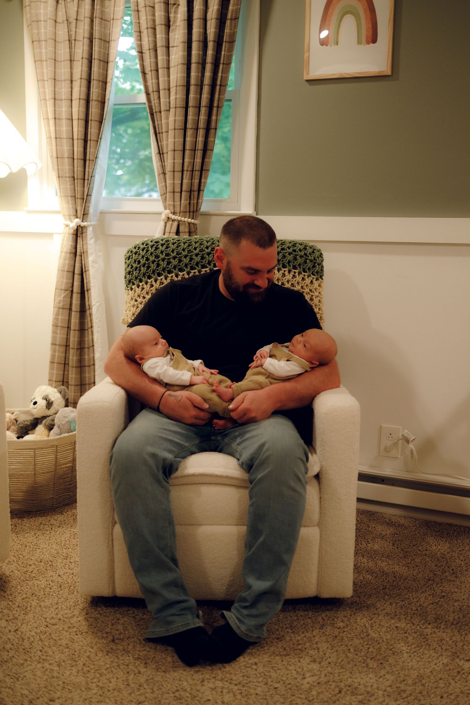 Someone sits in a white nursery chair holding two sleeping newborn twins while stuffed animals rest nearby in a basket.