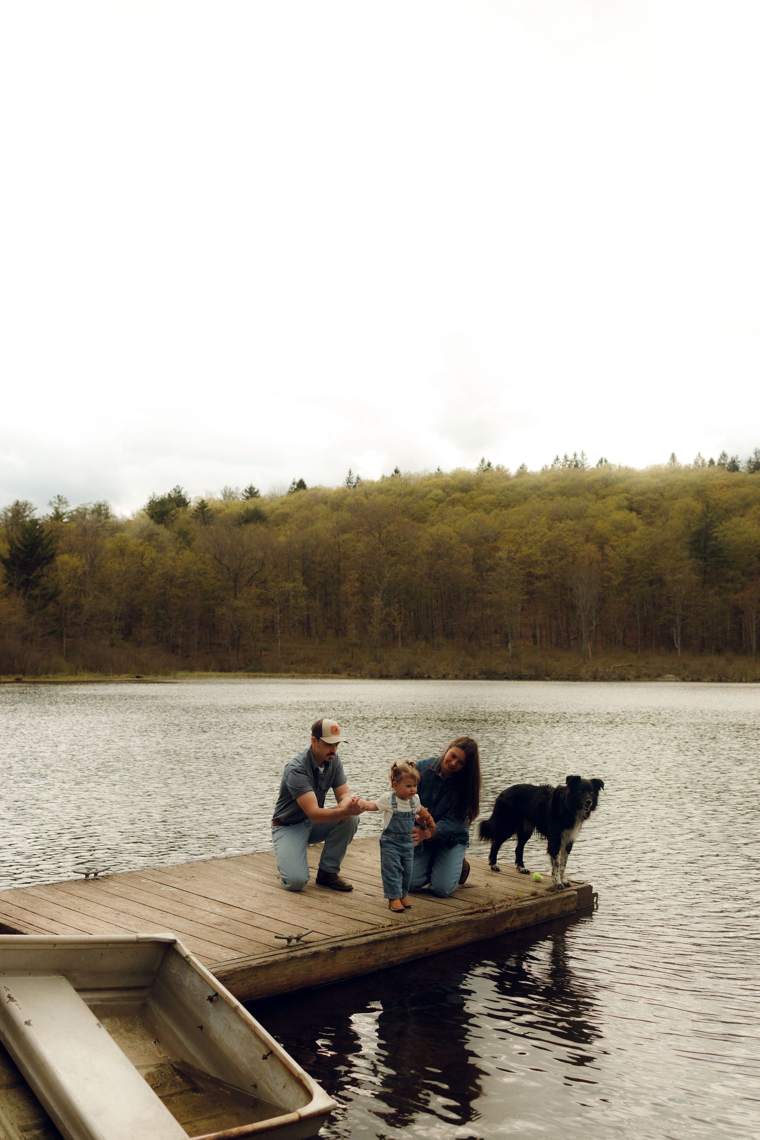 Two people sitting with their dog on a wooden dock by a peaceful lake surrounded by wooded hills on an autumn day.