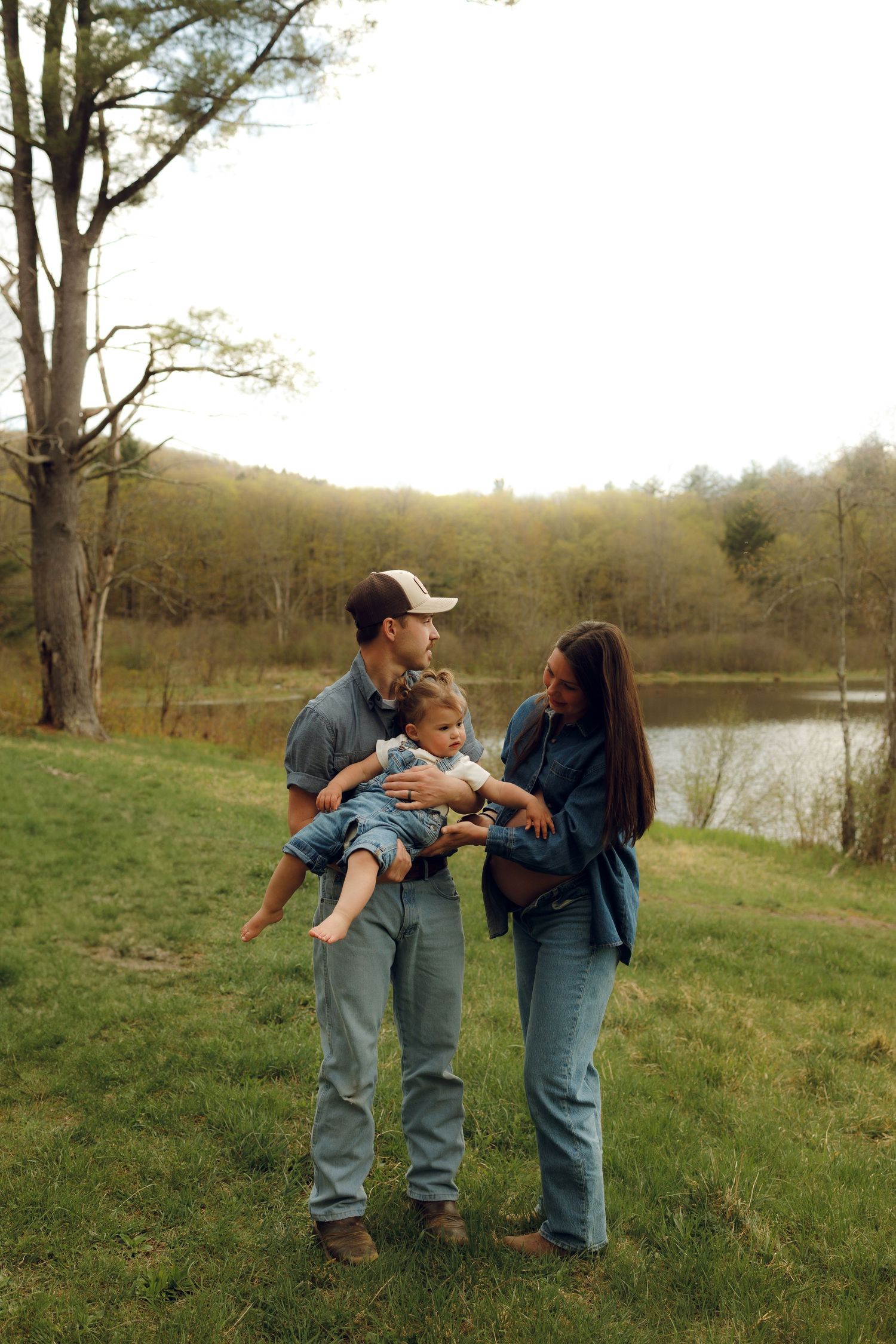 Three people stand together on a grassy hillside near a tranquil pond at sunset in a serene countryside setting.