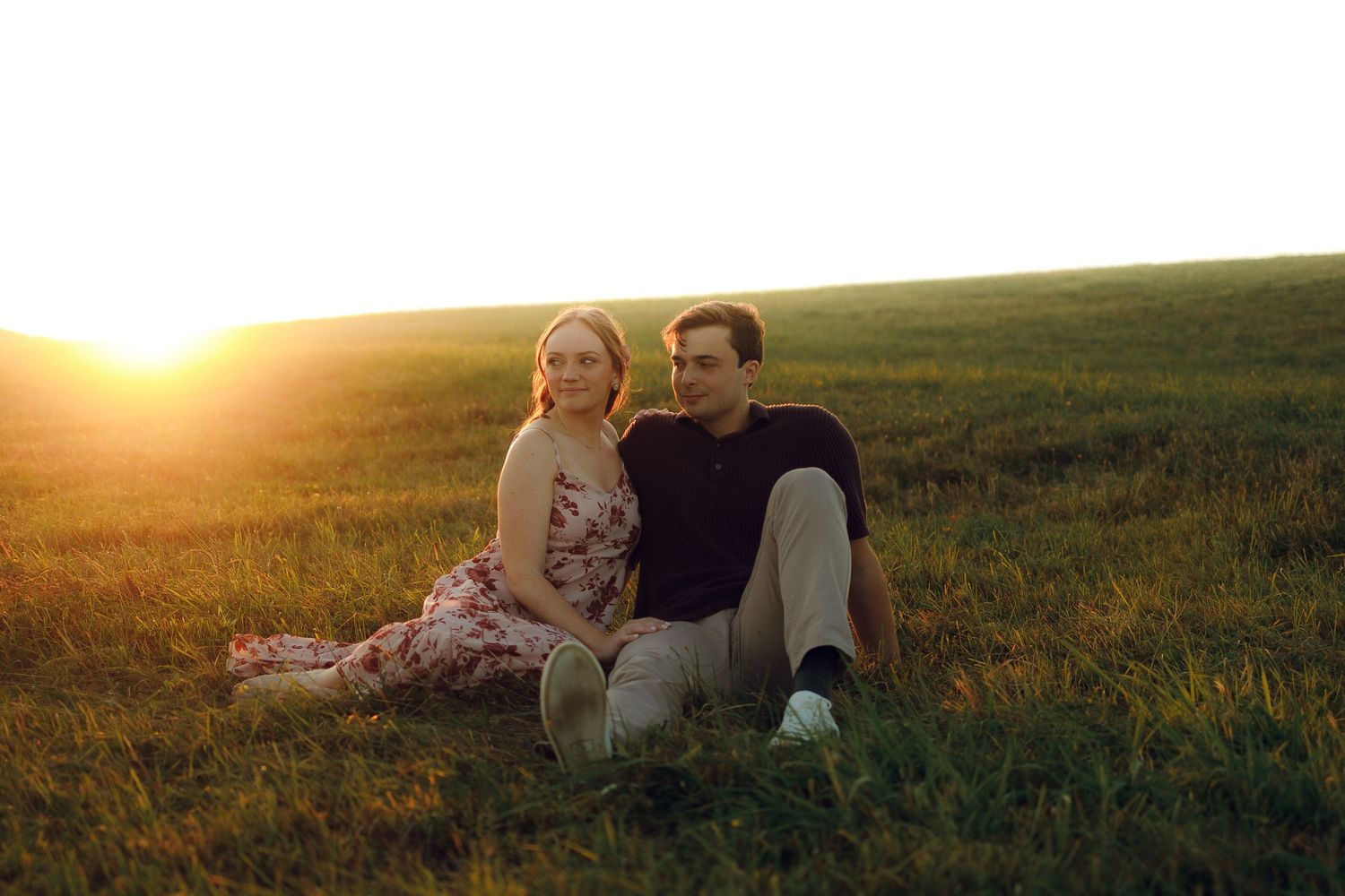 Couple embraces in a romantic sunset moment on a grassy hillside during a golden hour photoshoot.