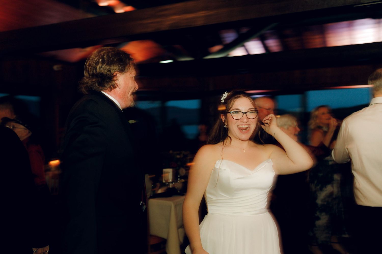 A bride in a white strapless dress and glasses dances joyfully at a wedding reception with motion blur in the background.