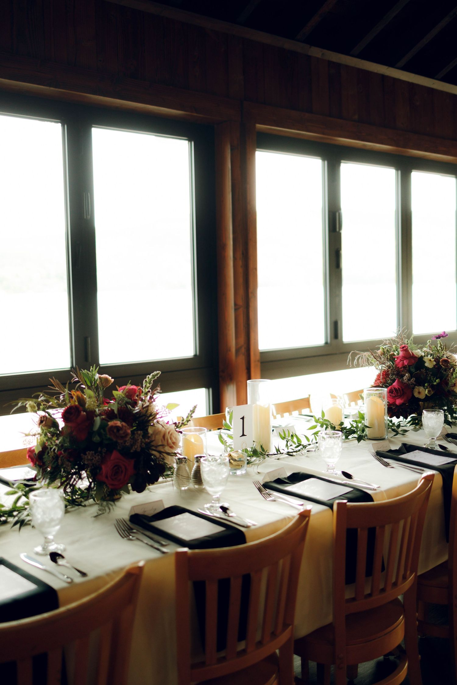 Elegant restaurant table settings with white linens and floral centerpieces line up along bright windows in a dining room.