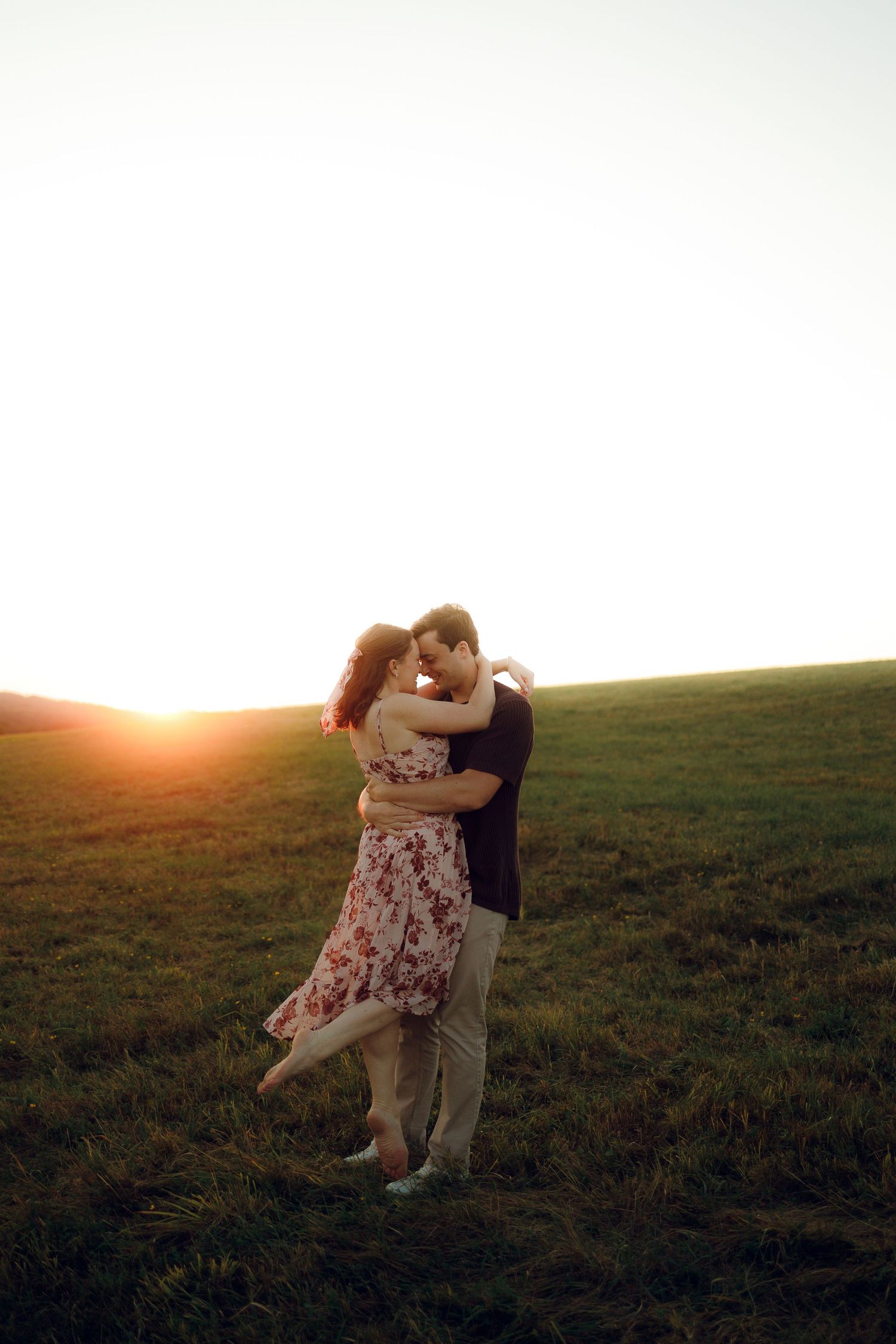 A couple embraces in a grassy field during golden hour sunset, creating a romantic silhouette against the glowing sky.