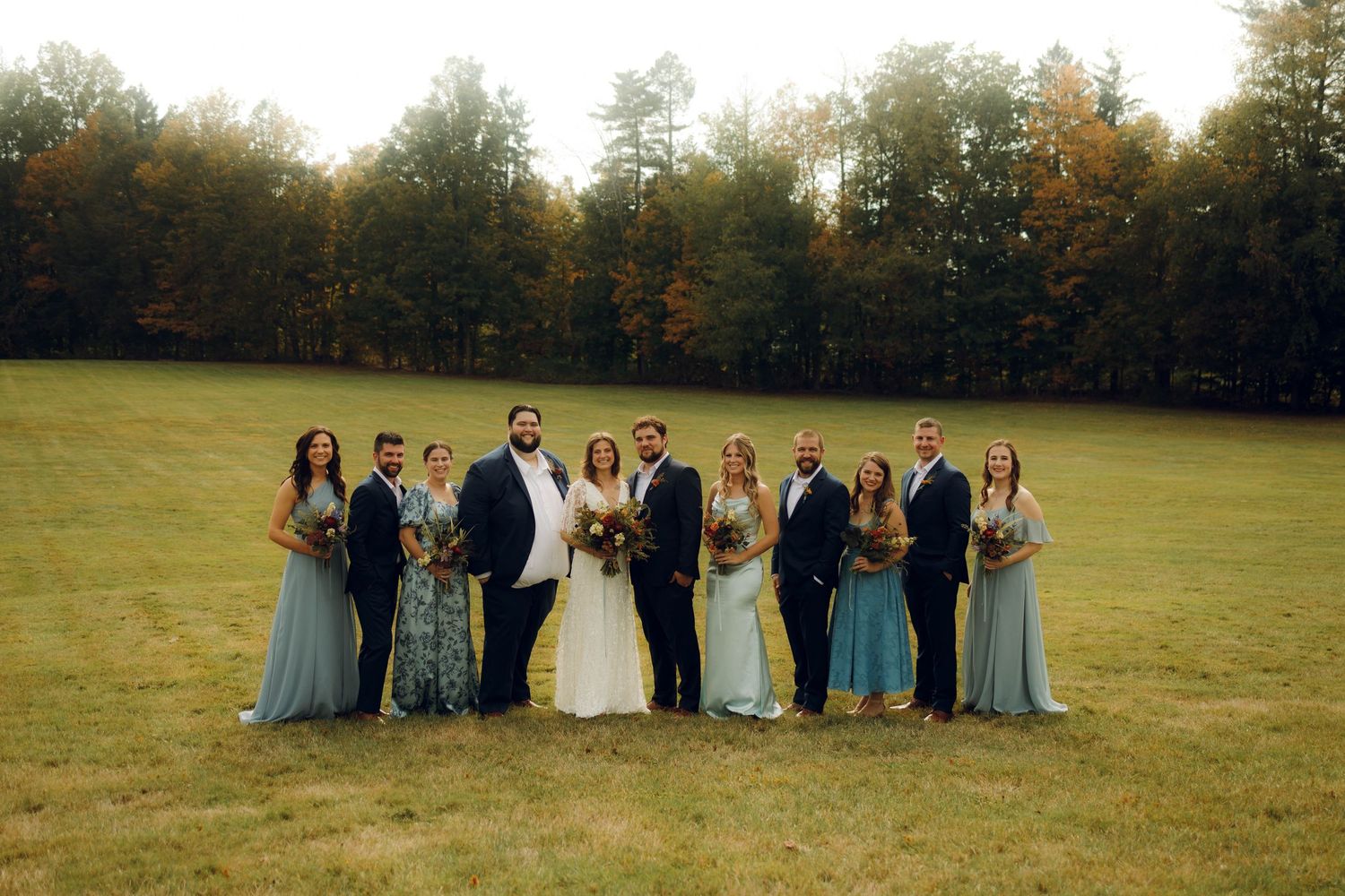 Wedding party poses in an autumn field at golden hour with warm sunlight filtering through trees in the background.